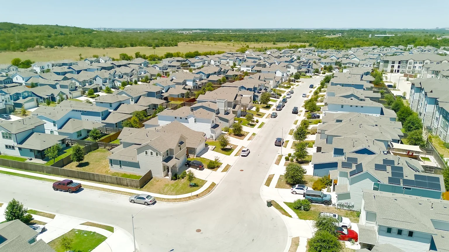 Aerial view of a suburban neighborhood with rows of houses, parked cars, green lawns, trees, and a central street.