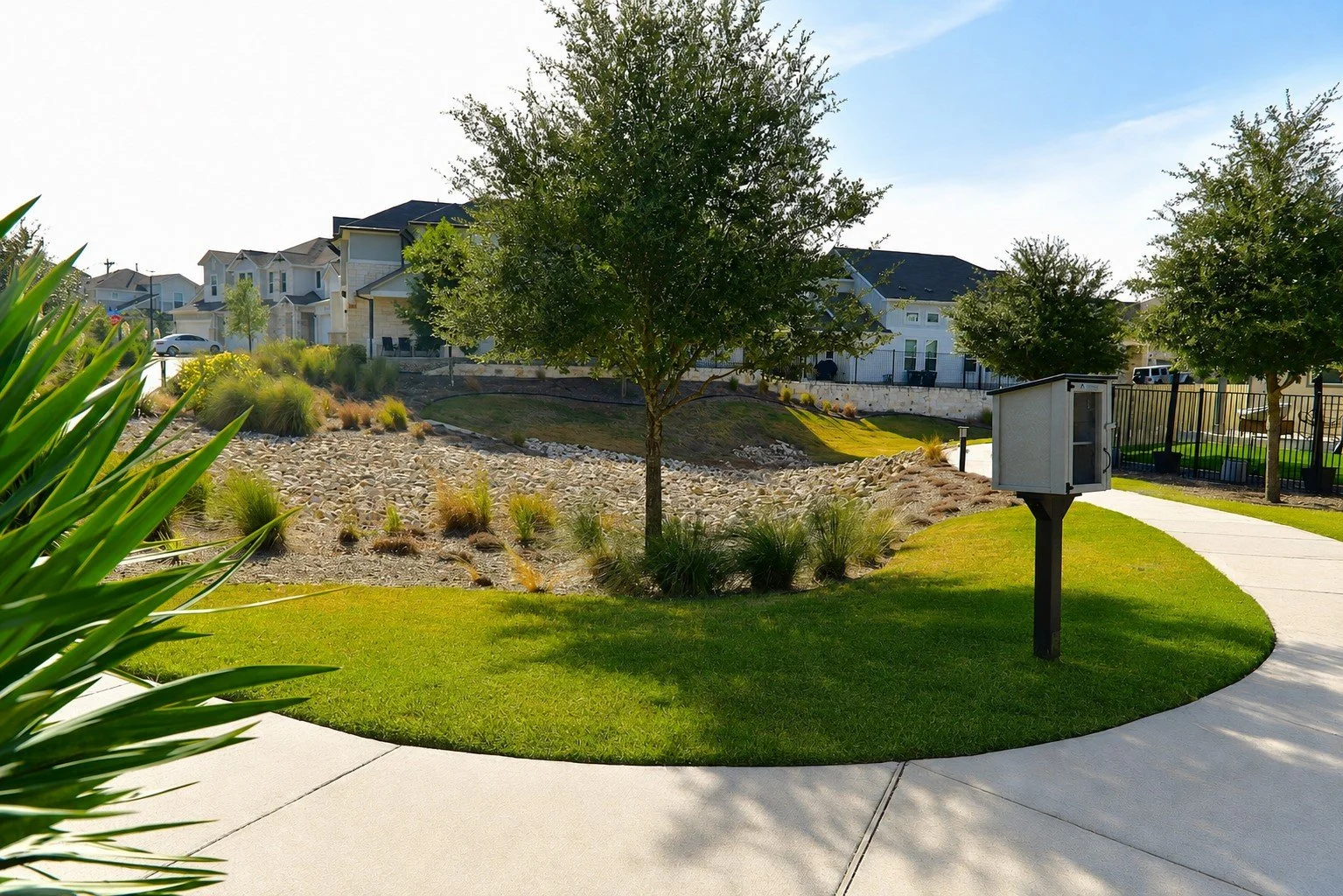 A landscaped park with a grassy area, a curved concrete walkway, a central tree, and decorative plants. There is a small white structure on a black post near the walkway. In the background, houses and a fenced playground are visible.