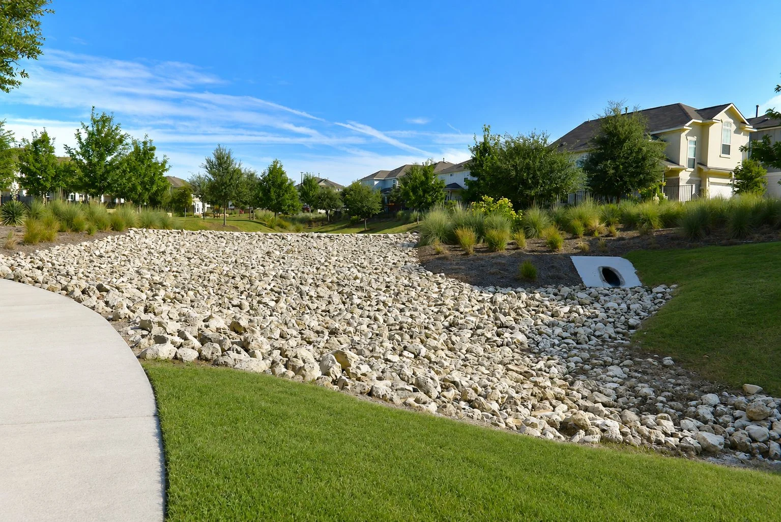 Residential neighborhood with well-maintained grass, sidewalk, and landscaping including trees, shrubs, and rocks with houses in the background under a blue sky.