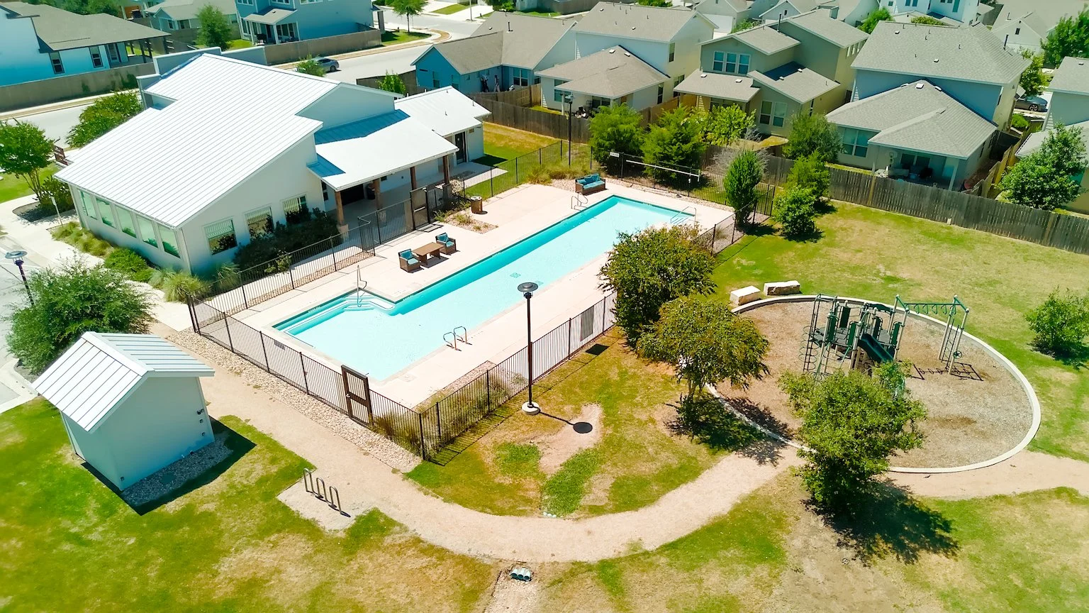 Aerial view of a community park featuring a swimming pool with surrounding patio, a small building, a children’s playground, and nearby residential houses.