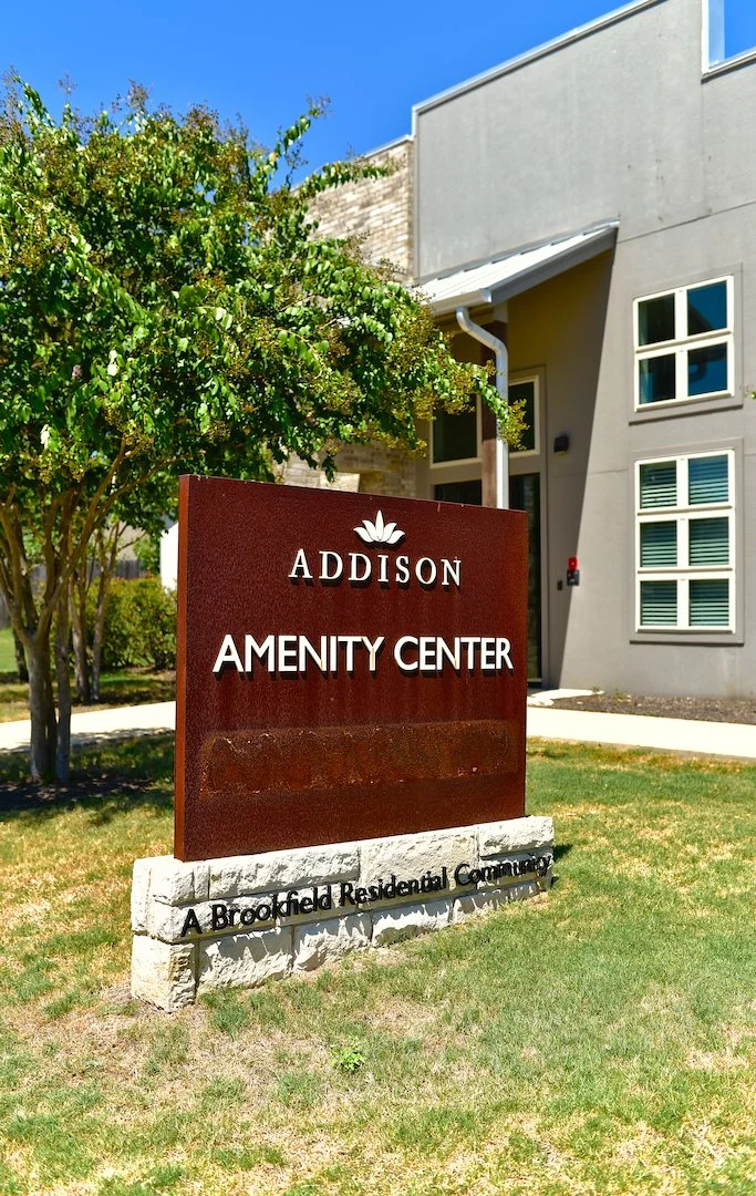 Rusty brown sign that reads 'ADDISON AMENITY CENTER' with a lotus flower logo on top and the phrase 'A Brookfield Residential Community' at the bottom, surrounded by green grass, trees, and a modern building in the background under a clear blue sky.