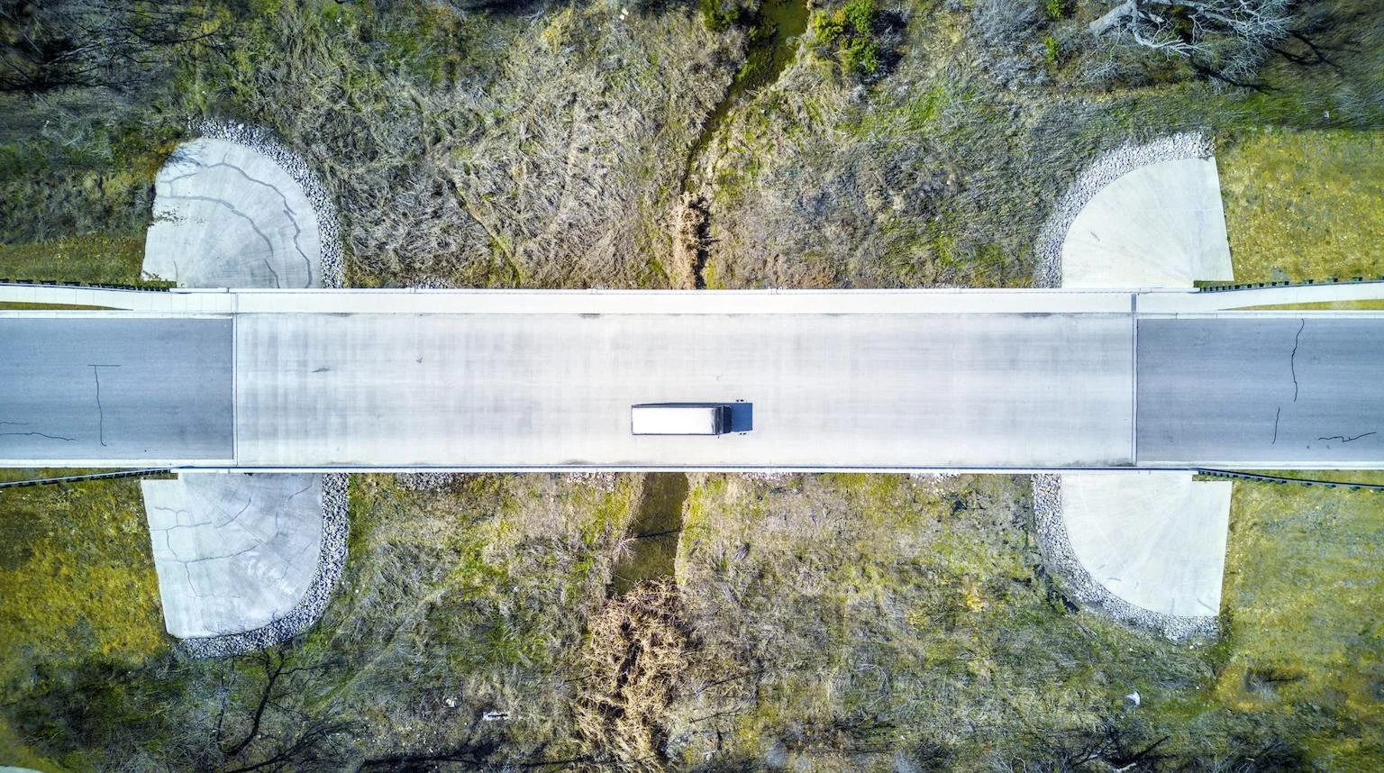 An aerial view of a bridge crossing over a small creek in a wooded area, with curved concrete supports on each end.