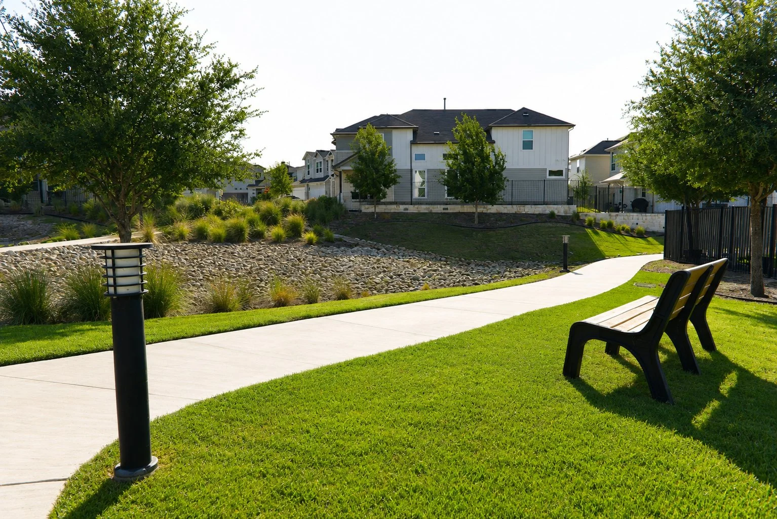 A park with a paved walking path, green grass, trees, and modern houses in the background. There are benches and a black fence along one side of the park.