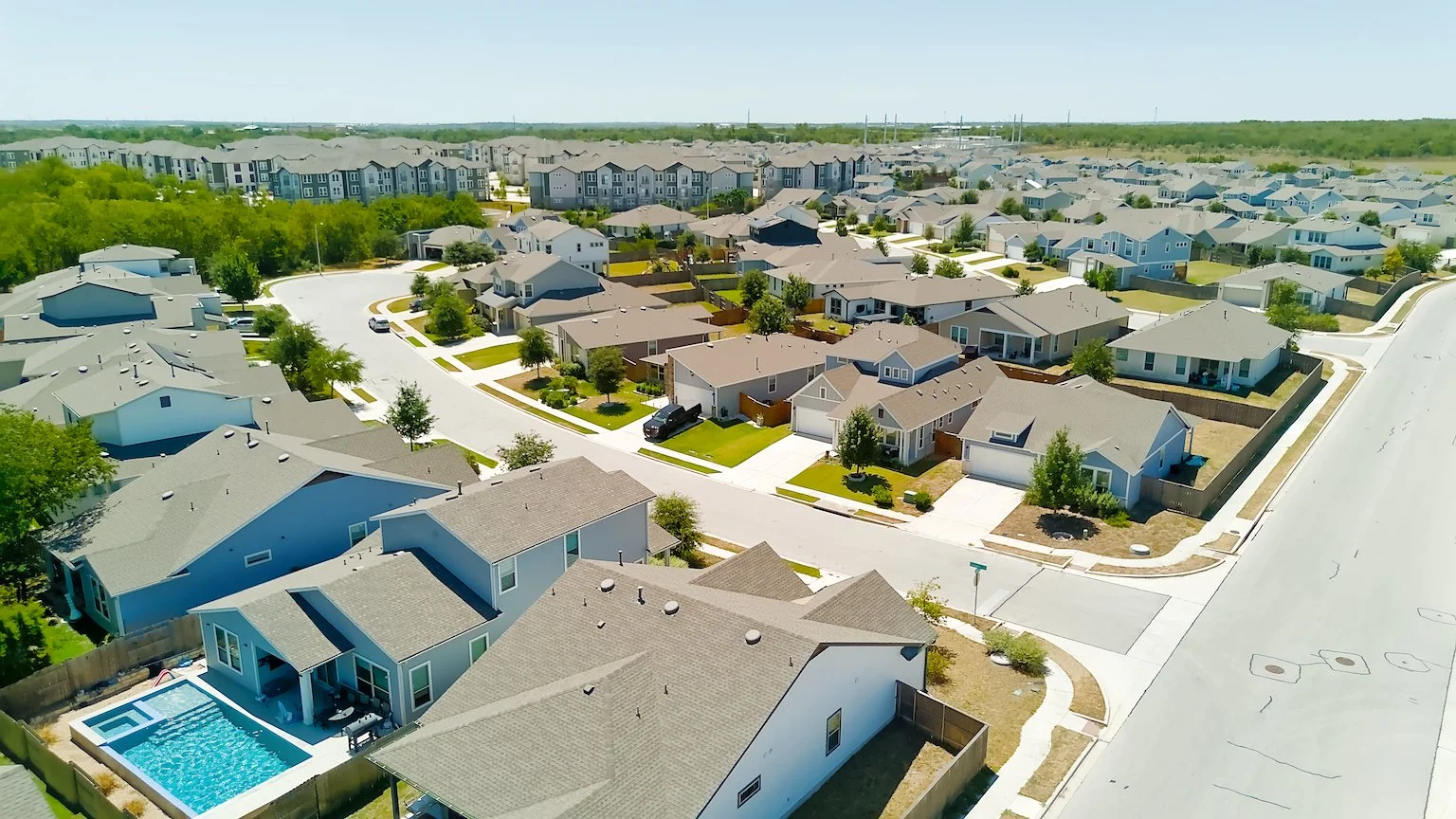 Aerial view of a suburban neighborhood with single-family homes, green lawns, trees, and a backyard with a swimming pool.
