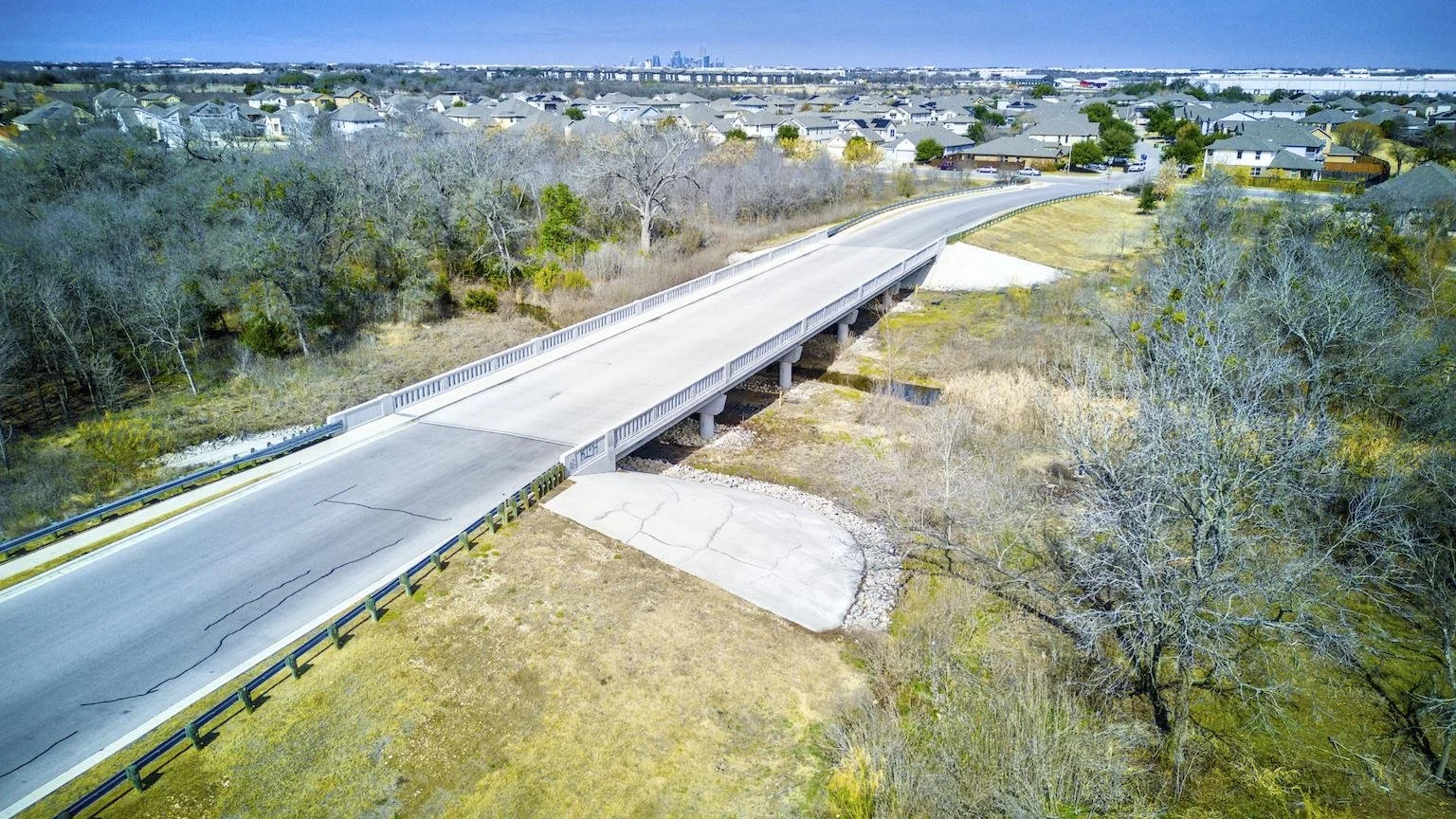 An aerial view of a concrete bridge over a grassy and wooded creek area leading to a residential neighborhood with houses and trees.