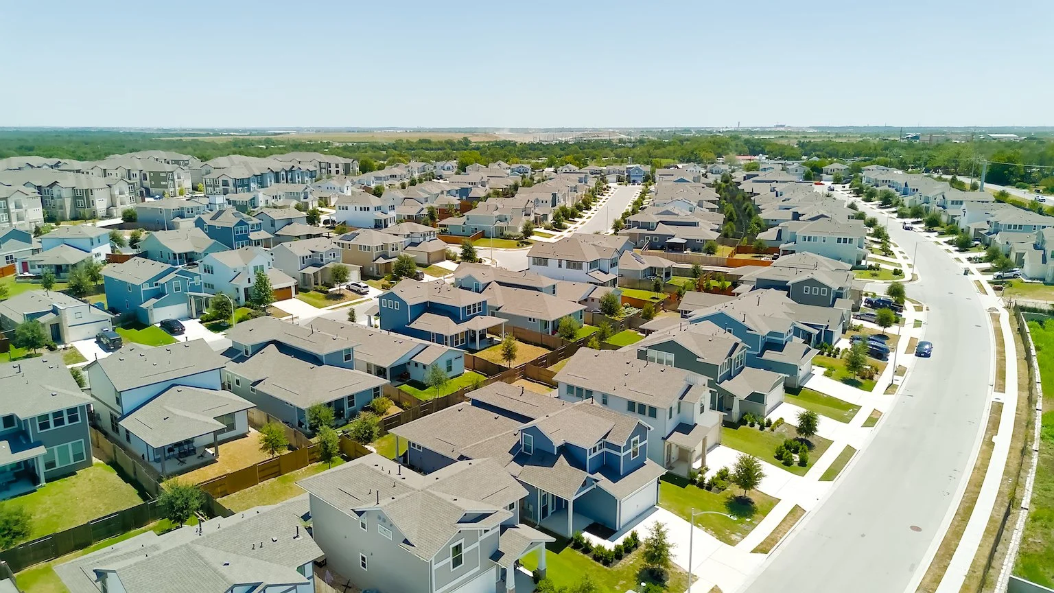 Aerial view of a suburban neighborhood with new houses, green lawns, and streets with cars under a clear blue sky.