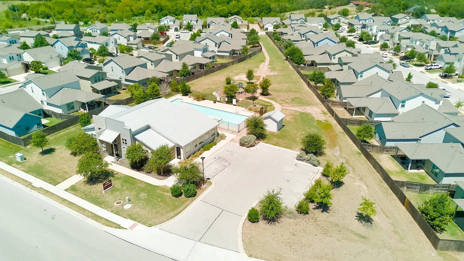 Aerial view of a community park with a swimming pool, basketball court, and playground, surrounded by residential houses with gray roofs.