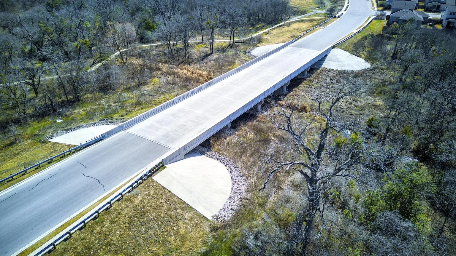 Aerial view of a concrete road bridge crossing over a creek with trees and grass.