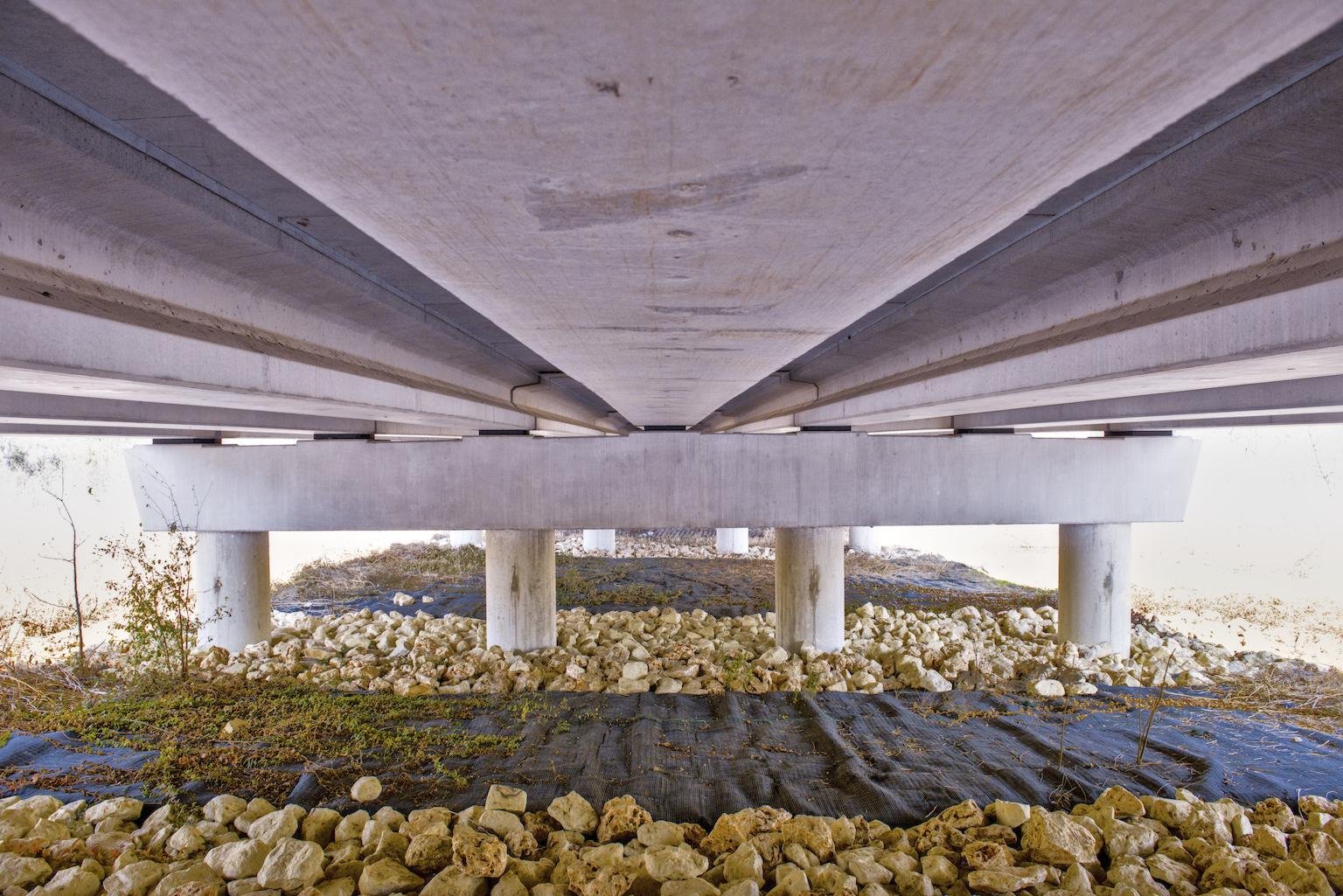 Underneath a bridge or overpass supported by large concrete pillars with gravel and some plants on the ground.