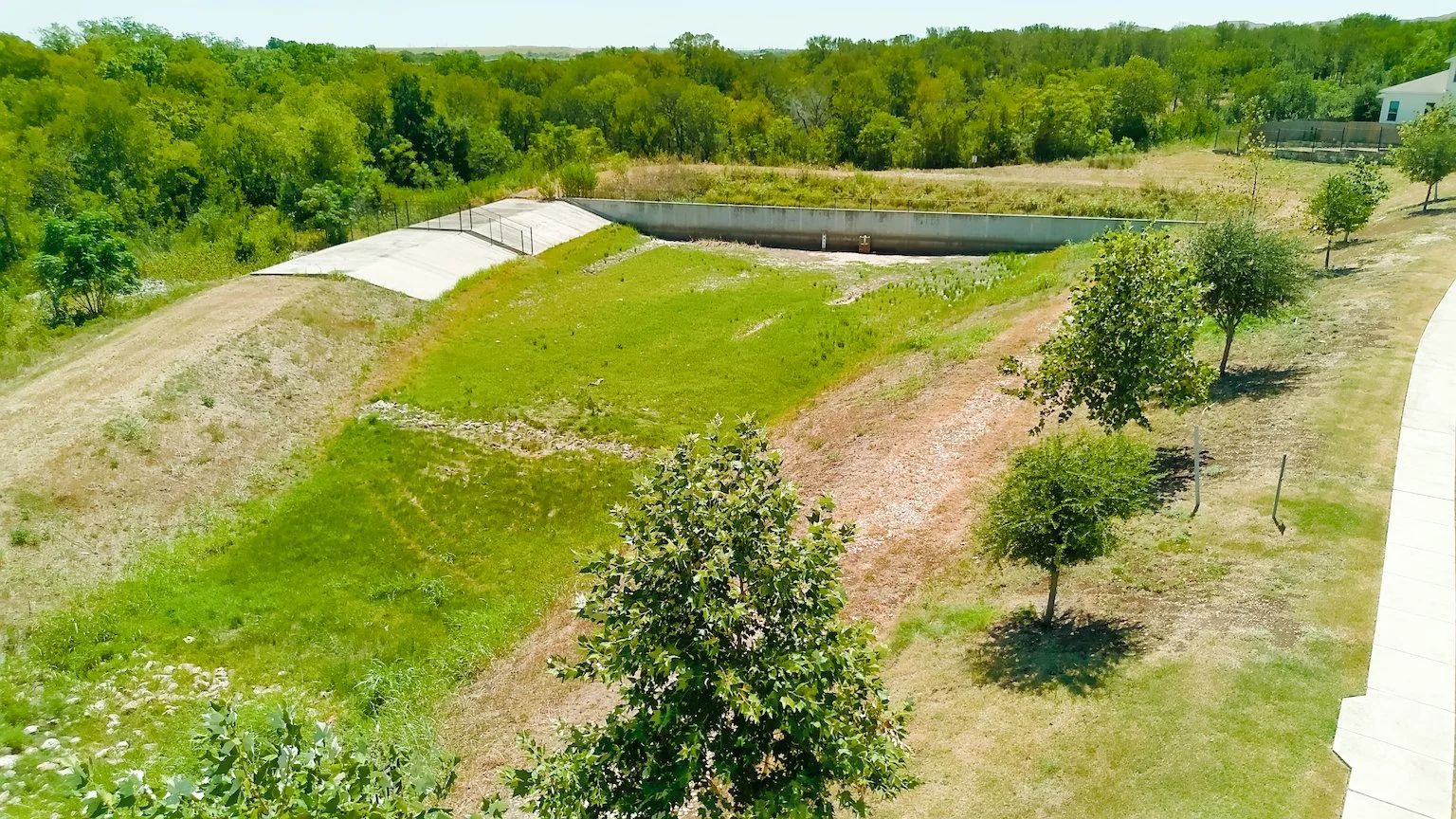 A view of a small pond or water retention area with a concrete lining, surrounded by grassy and wooded areas, and a paved walkway alongside the grassy area.