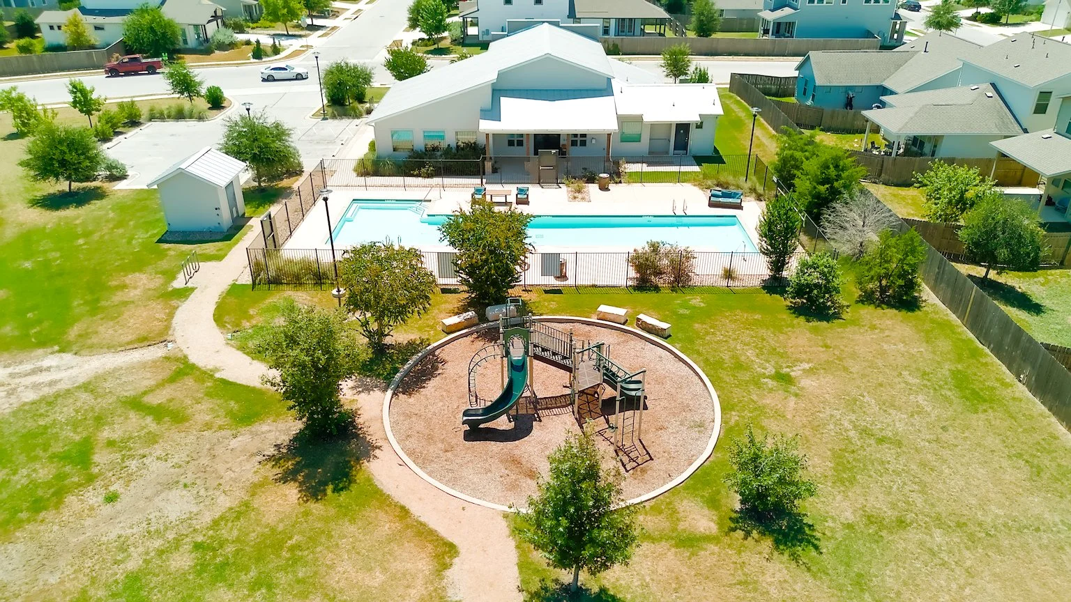 Aerial view of a community park with playground equipment, a picnic area, and a swimming pool. Surrounding residential houses and streets are visible.