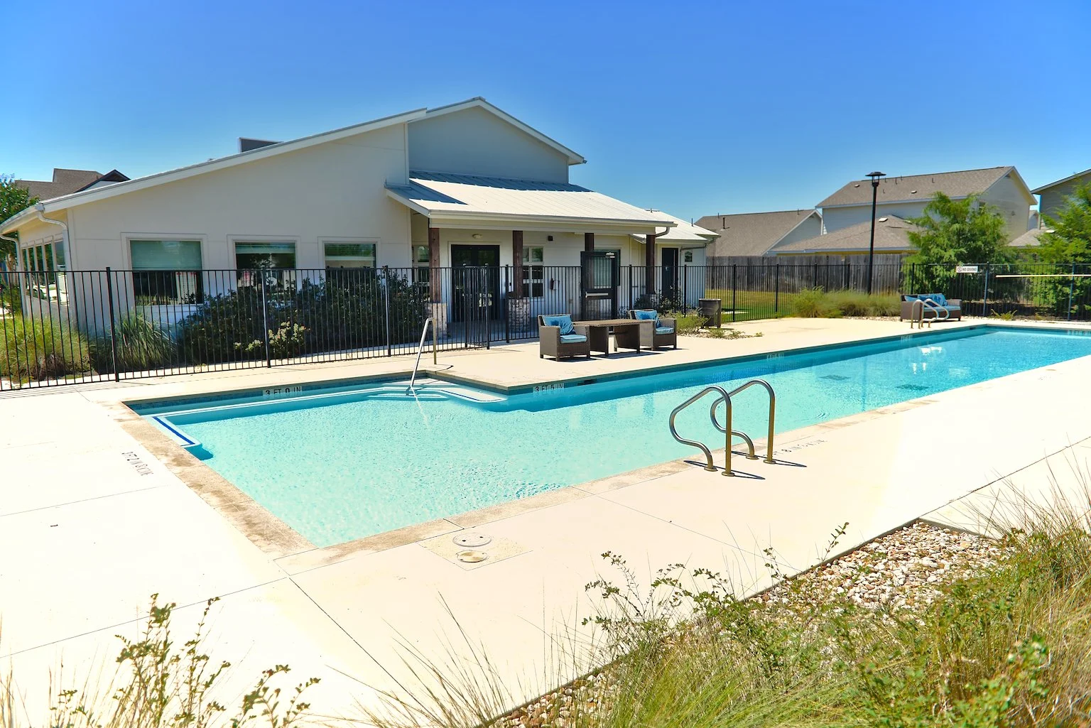 Residential backyard with a swimming pool, patio furniture, and a white house in the background on a sunny day.