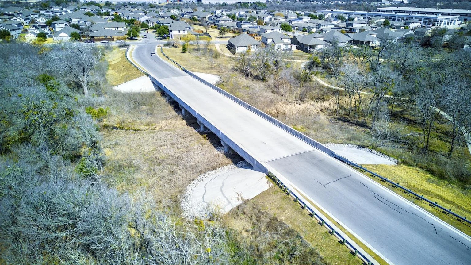 An aerial view of a newly constructed concrete bridge over a grassy and wooded creek area, leading into a residential neighborhood with numerous houses and streets.
