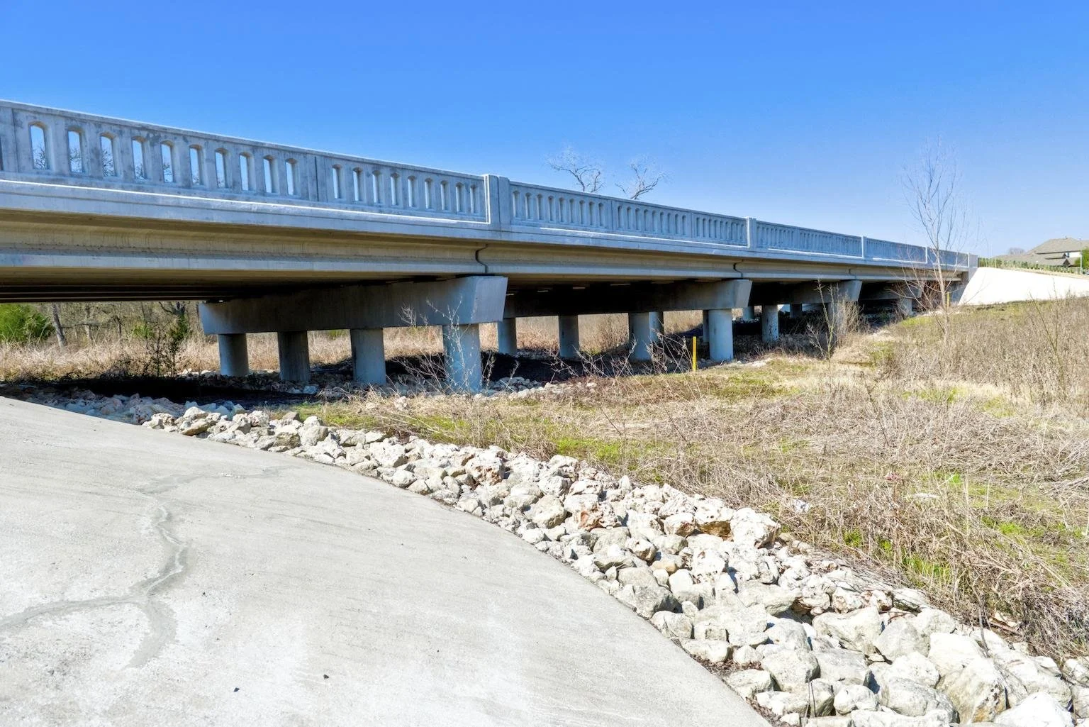 A bridge with concrete supports over dry, grassy land under a clear blue sky.