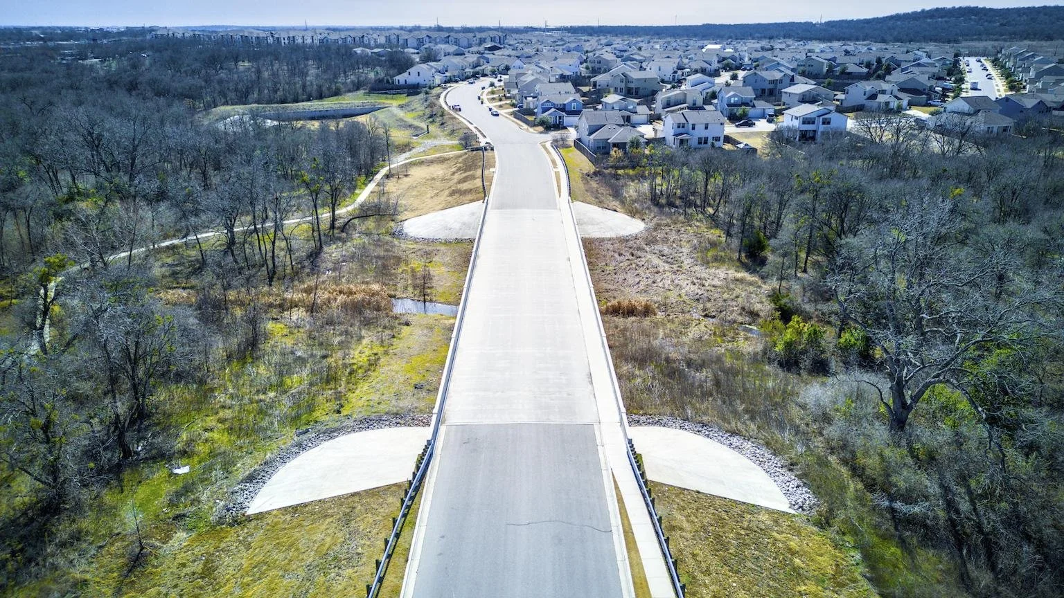 An aerial view of a newly constructed bridge extending over creek towards a suburban neighborhood with houses and streets.