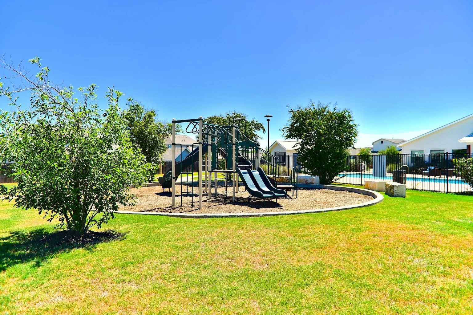 A playground with metal slides and climbing structures surrounded by a fence, with a swimming pool and residential houses in the background under a clear blue sky.