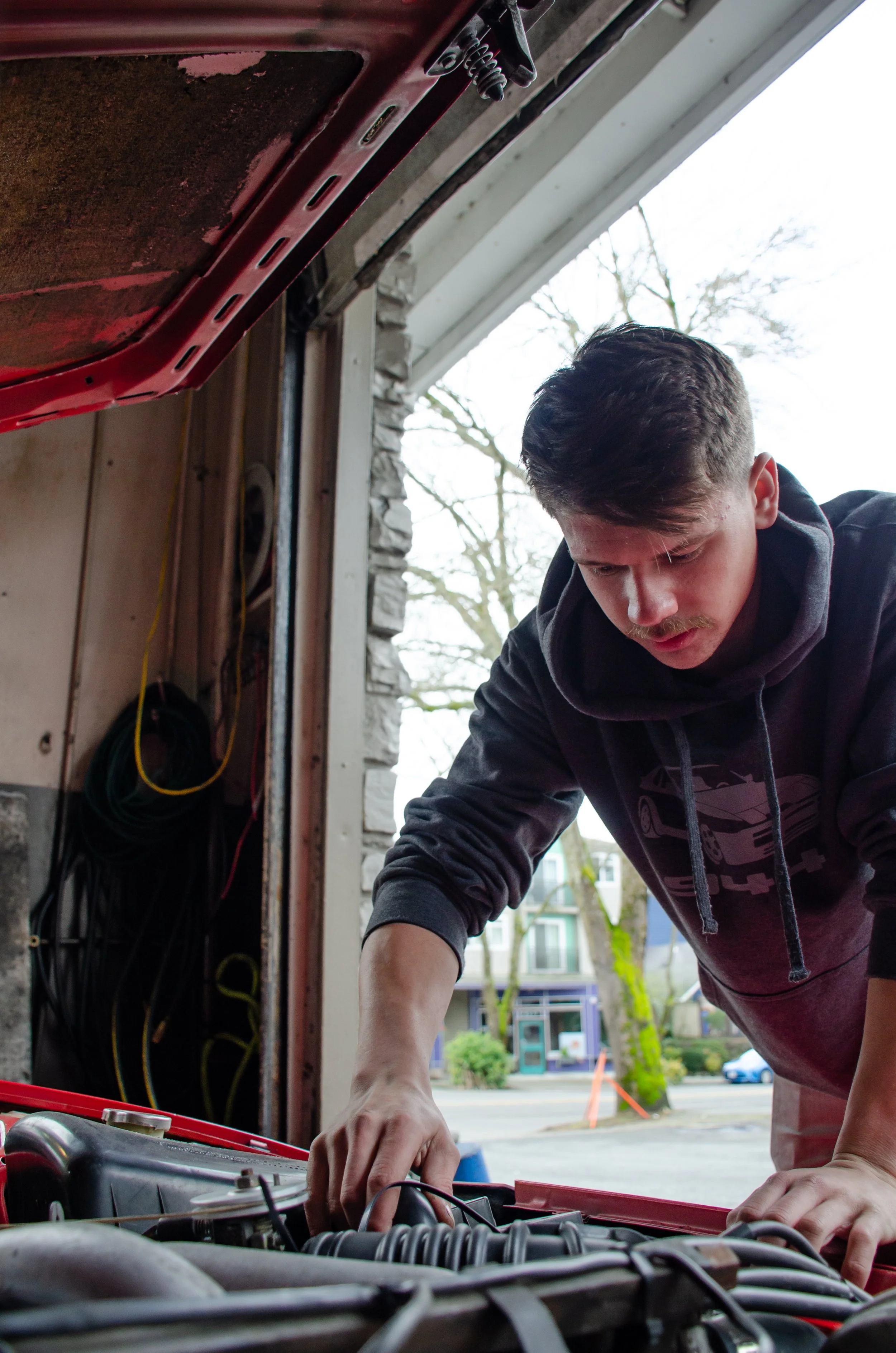 A young man working on a car engine in a garage, with his hand reaching into the engine bay. Outside view through the open garage door shows a street with trees and buildings.