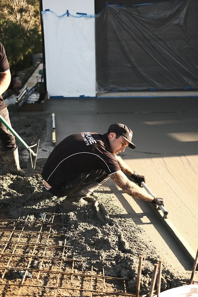 Construction worker smoothing wet concrete on a sidewalk using a tool, with rebar and construction materials visible nearby.