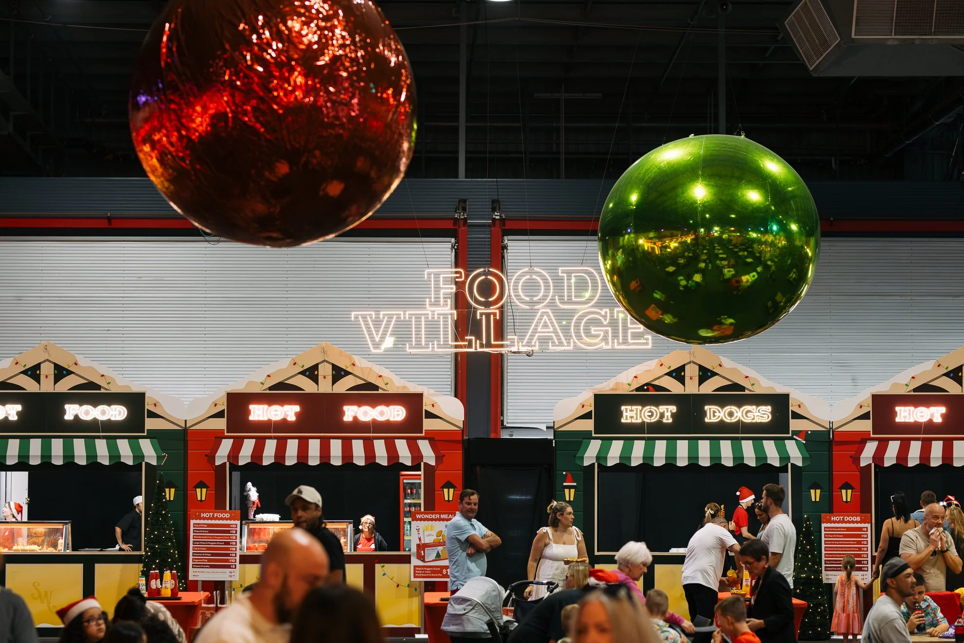Holiday-themed food stall at a food court with a neon sign that reads 'Food Village' and decorative ornaments hanging from the ceiling, including large red and green reflective spheres.