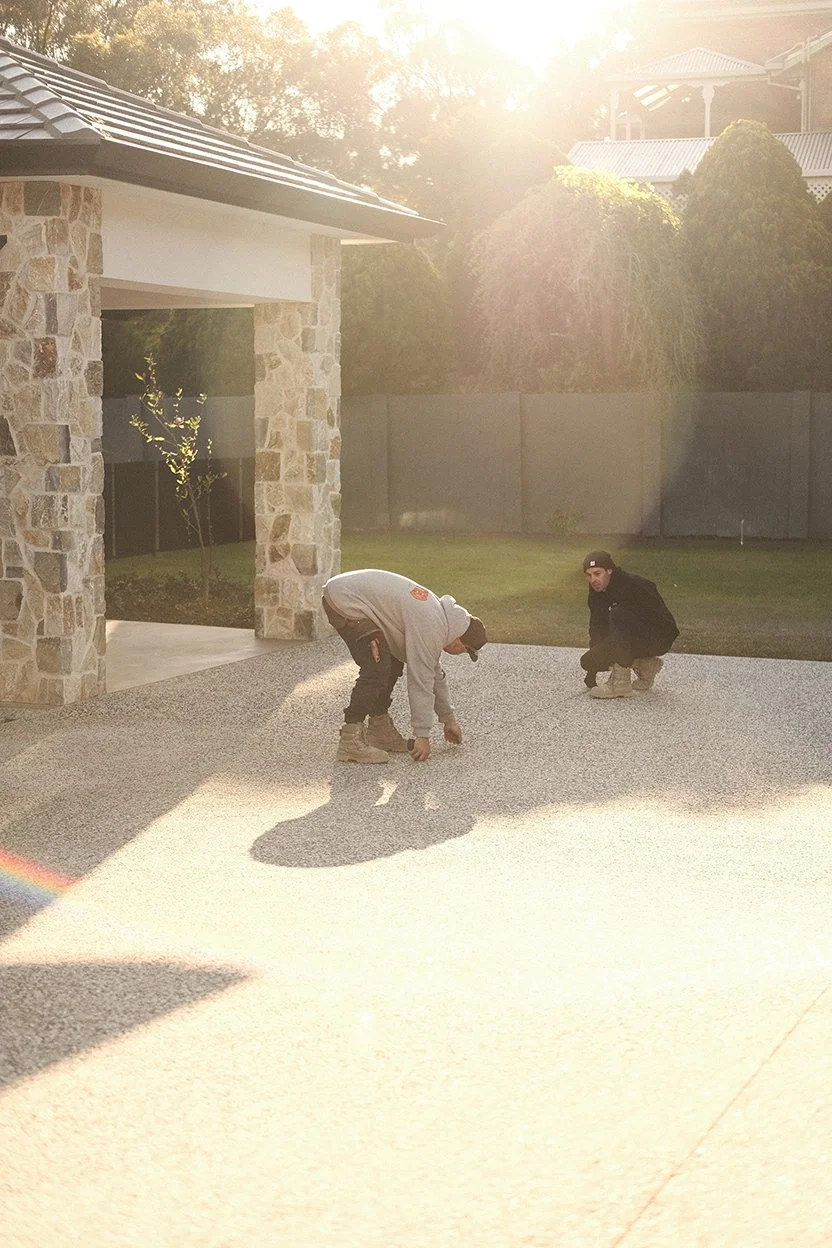 Two men working on a gravel driveway outside a house with stone columns, trees, and a fence, sun shining brightly in the background.