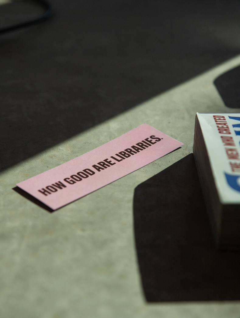 Pink sign with black text that says "How good are libraries" placed on a gray surface, with part of a box of tissues visible on the right.