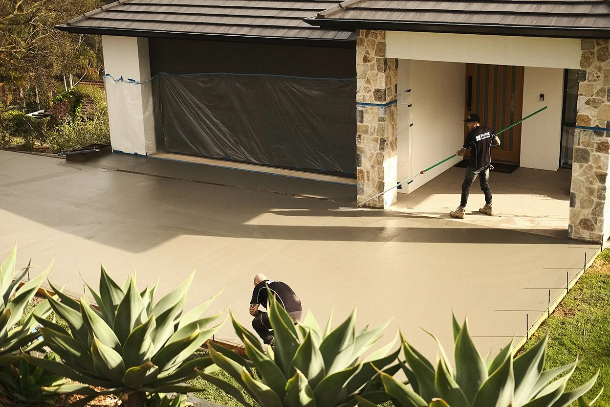 Two construction workers working on a new concrete driveway in front of a modern house with stone and white exterior walls.