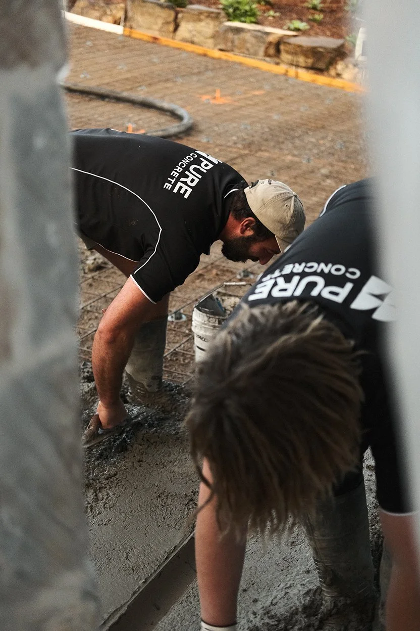 Two workers in black shirts and beige caps pouring and spreading wet concrete on a construction site.