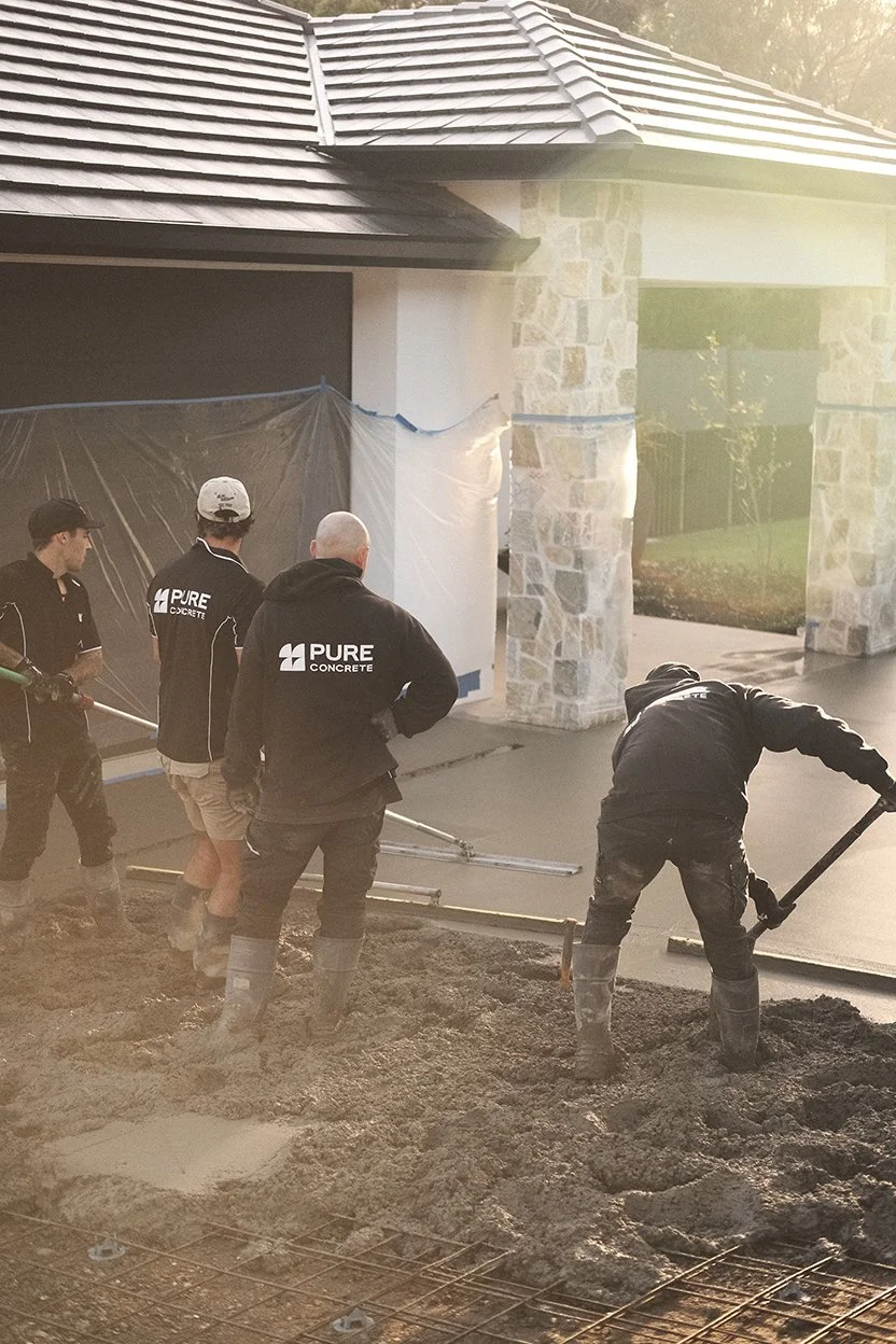 Construction workers leveling and smoothing freshly poured concrete in front of a house with stone pillars.