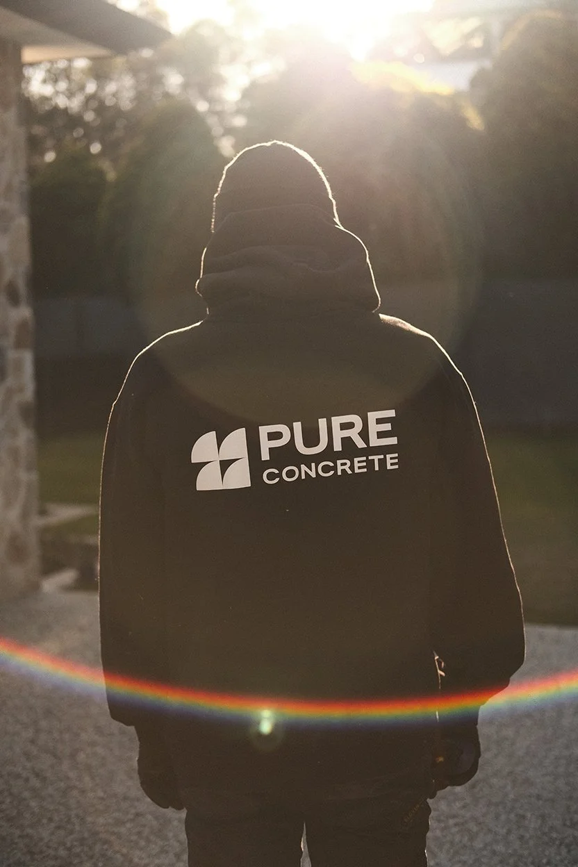 Person standing outdoors at sunset, wearing a black hoodie with a logo that says 'PURE CONCRETE' on the back.