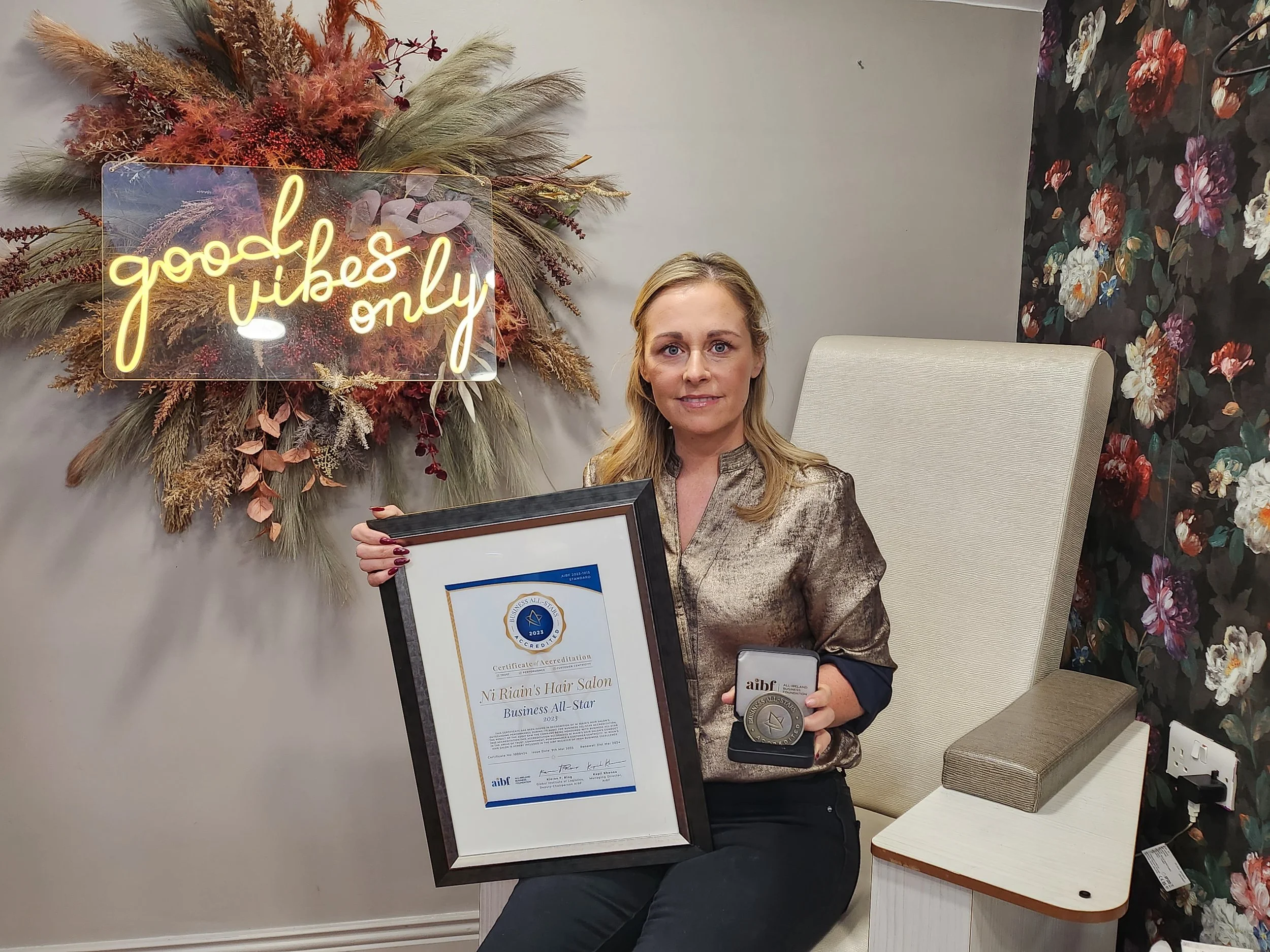 A woman sitting in a floral wallpapered room holding a certificate and a medal. Behind her is a decorative wall arrangement with dried plants and a neon sign that reads "good vibes only."