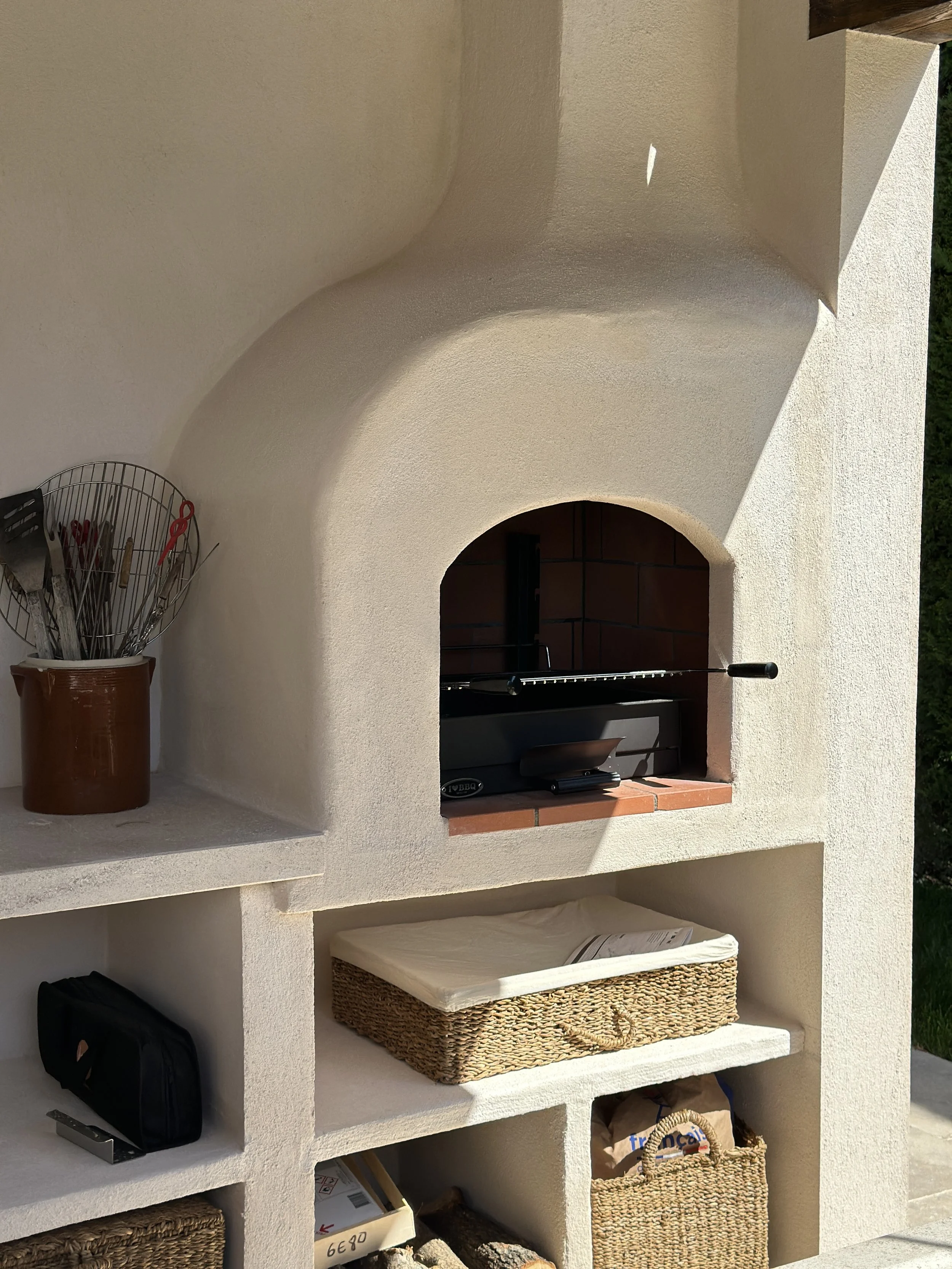 Cuisine d'été en Provence : barbecue maçonné à l'enduit clair, niches de rangement, paniers tressés et pot en terre cuite.