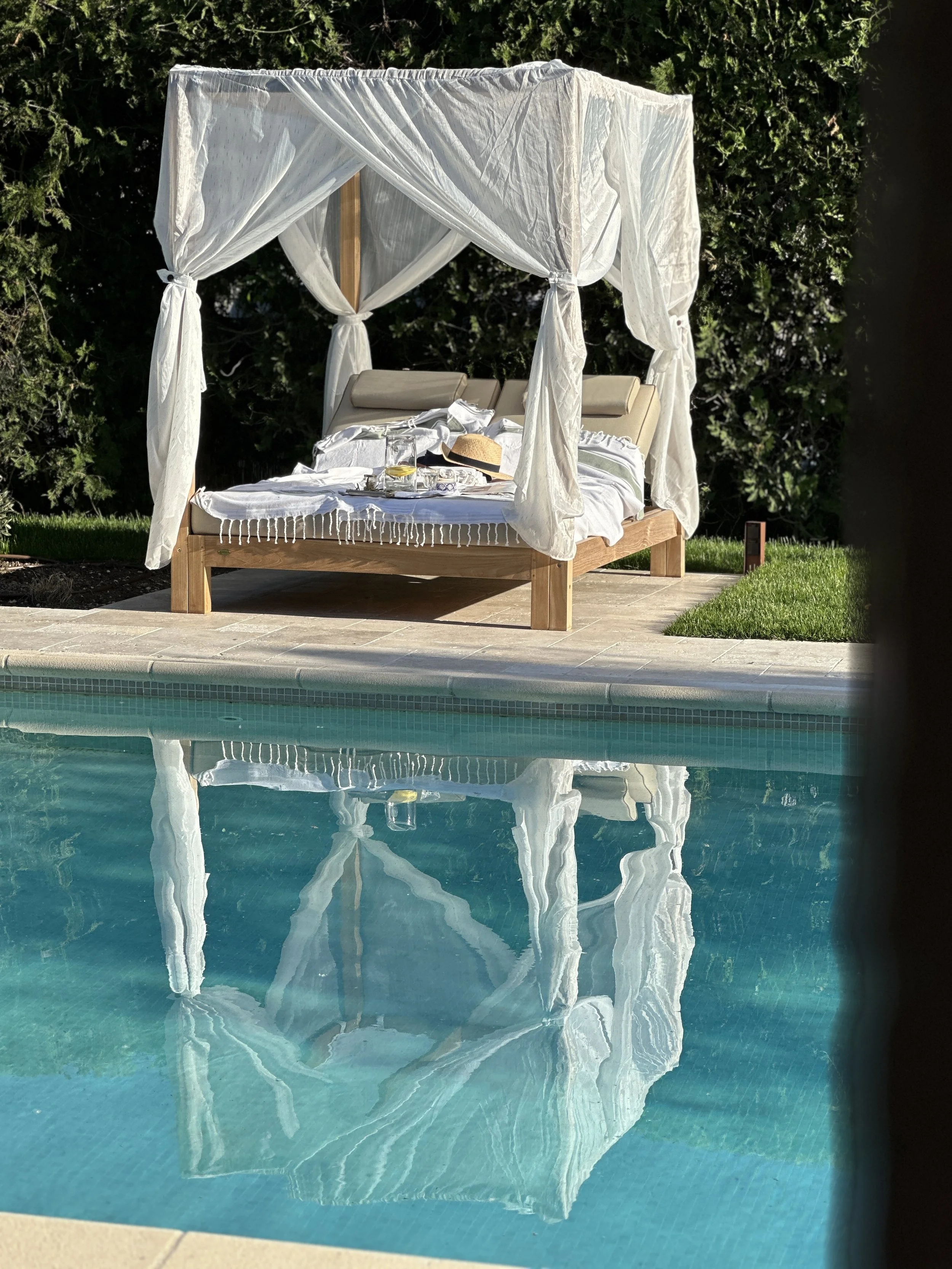 Lit à baldaquin en bois avec voilages blancs au bord d'une piscine turquoise, reflet dans l'eau et jardin verdoyant