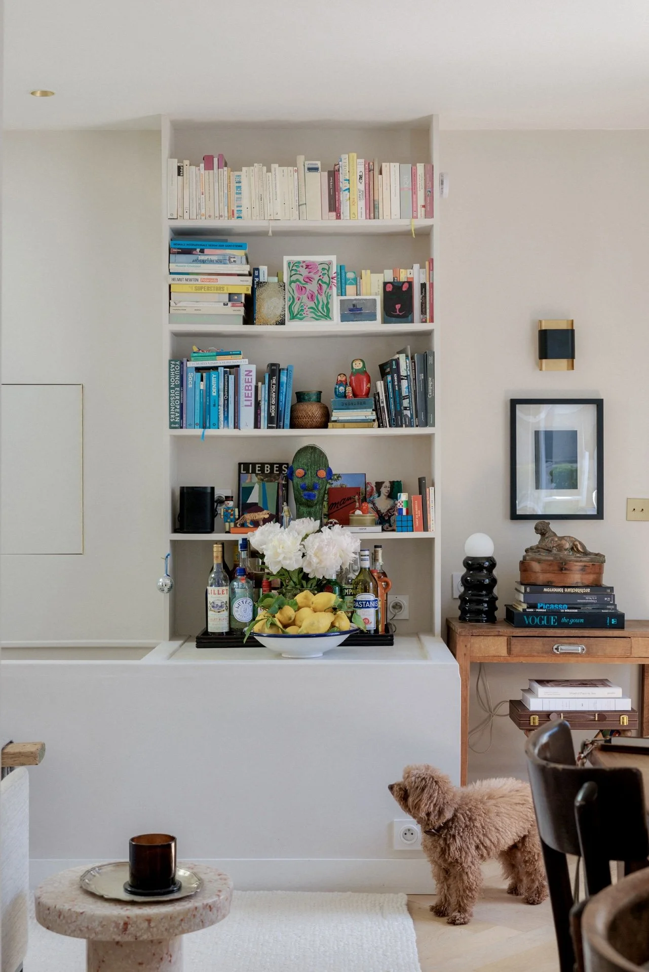 Salon avec bibliothèque murale intégrée remplie de livres et objets décoratifs, bar avec citrons et pivoines, chien caniche et console en bois