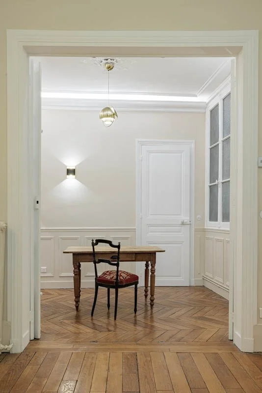 Bureau ancien chiné et chaise en bois noirci à assise brodée, parquet en point de Hongrie et moulures d'origine, sous une suspension en laiton doré.