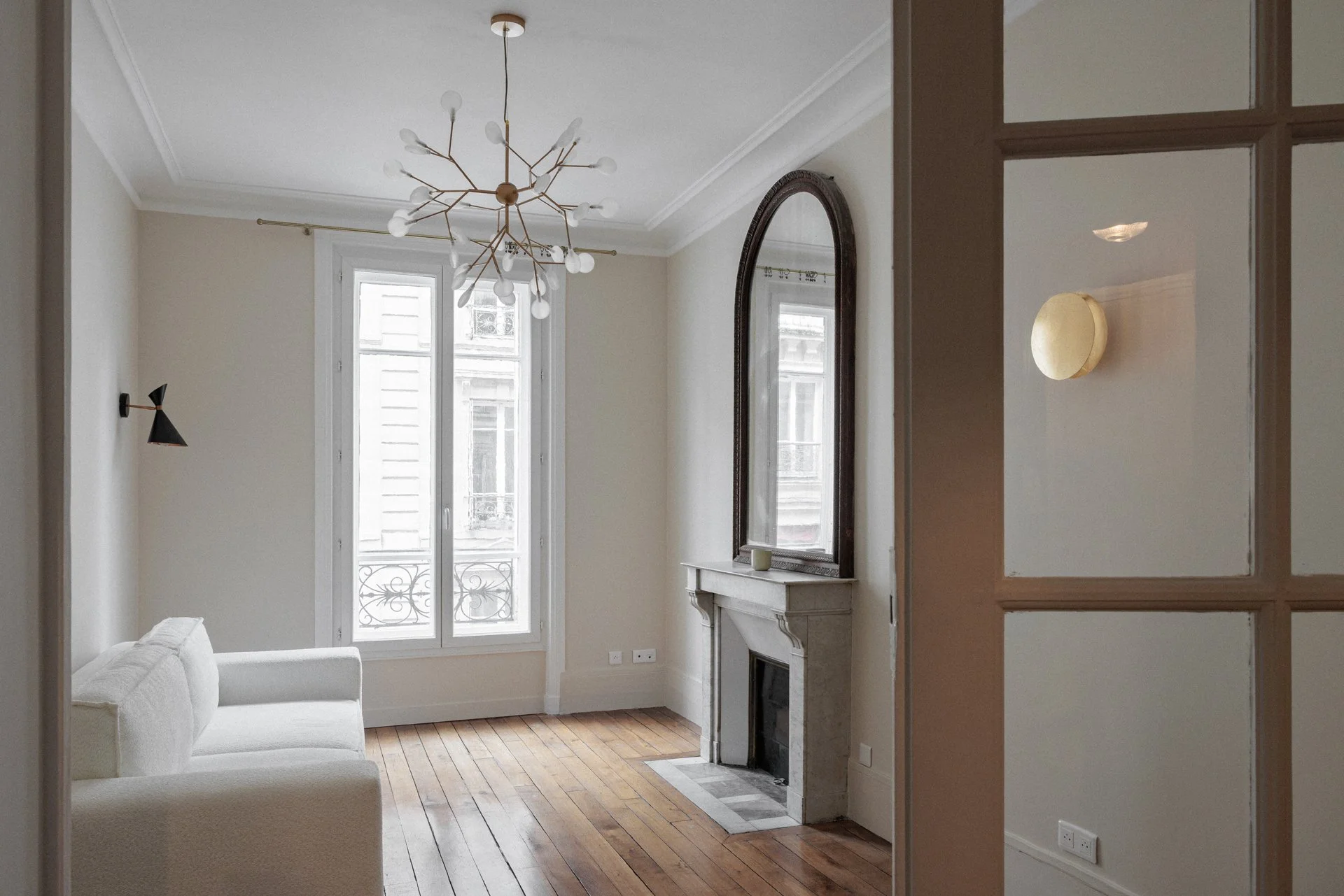 Salon parisien avec cheminée en marbre d'origine, grand miroir ancien à cadre bois, lustre contemporain en laiton et verre soufflé, parquet à bâtons rompus et porte vitrée en laiton donnant sur un balcon filant.