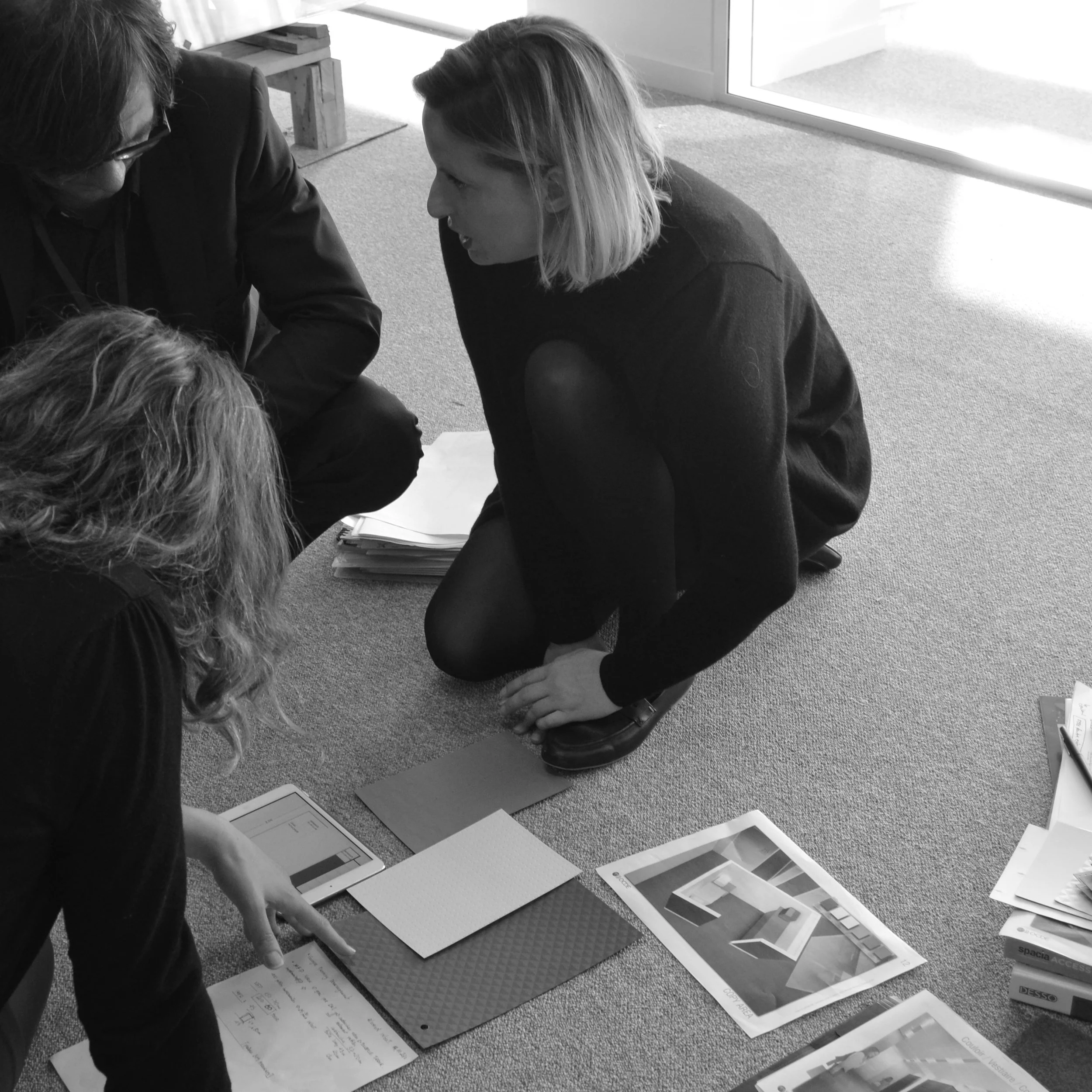Photo en noir et blanc d'une réunion de travail, l'équipe agenouillée au sol comparant échantillons de moquette et de sol, planches de rendus pour l'aménagement des bureaux OCDE Boulogne