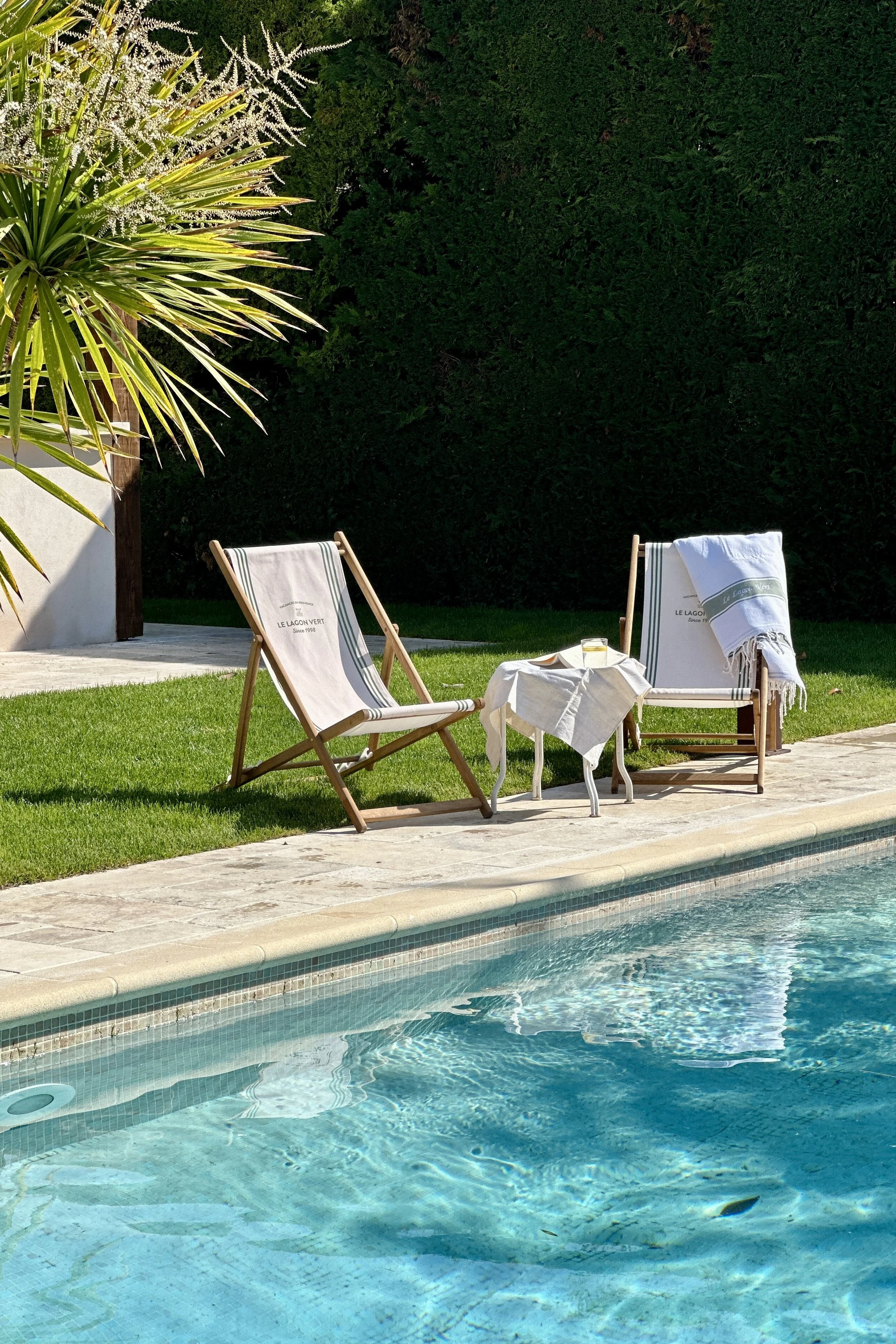 Deux transats en bois avec serviettes rayées au bord d'une piscine turquoise, petite table et palmier en arrière-plan