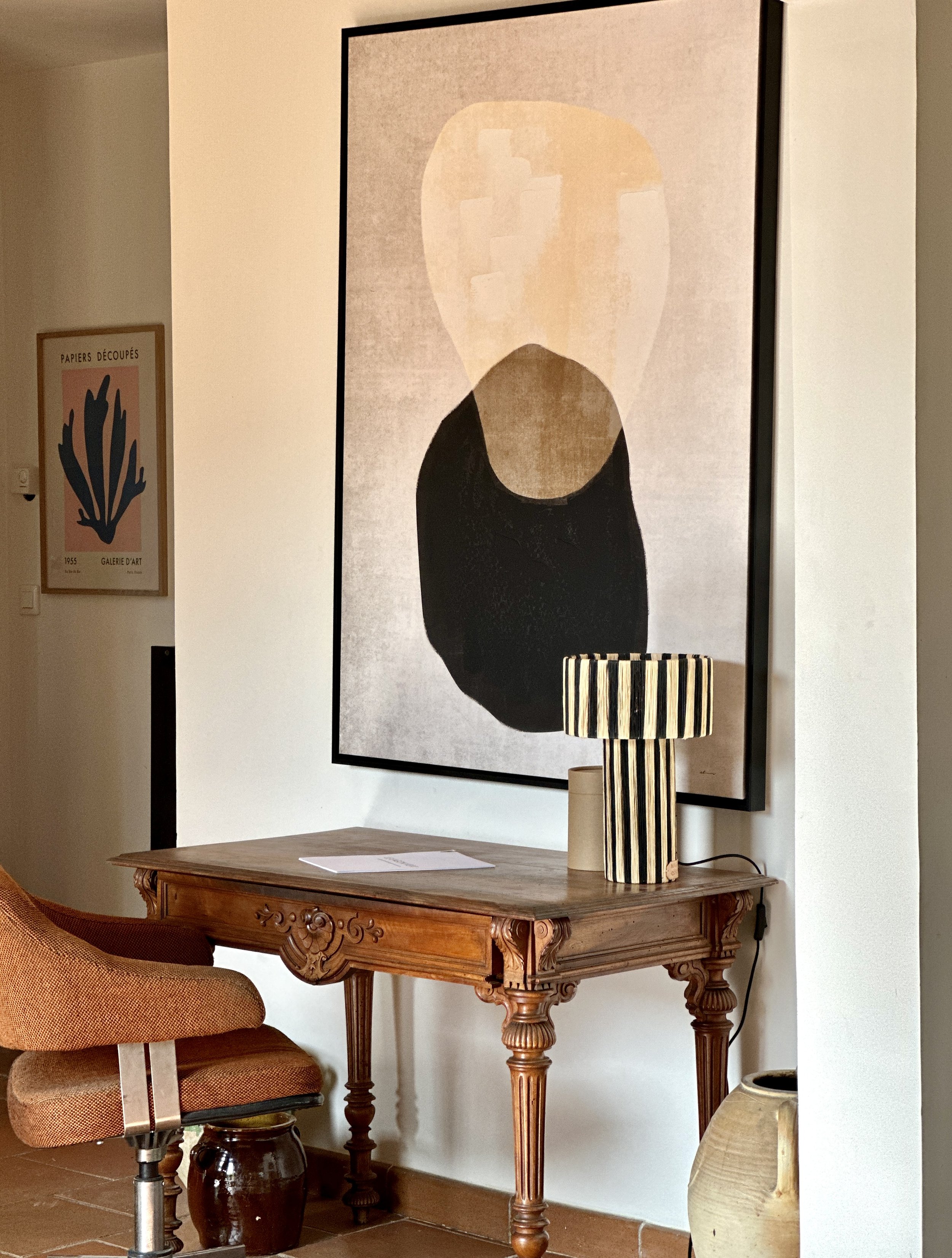 Console ancienne en bois sculpté avec lampe rayée noir et blanc, sous grand tableau abstrait, fauteuil vintage et jarres en terre cuite
