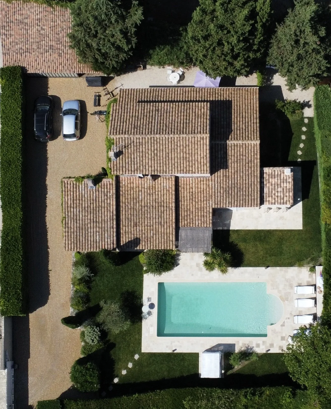 Vue aérienne de maison de campagne avec toit en tuiles, piscine turquoise, jardin arboré et allée avec voitures garées