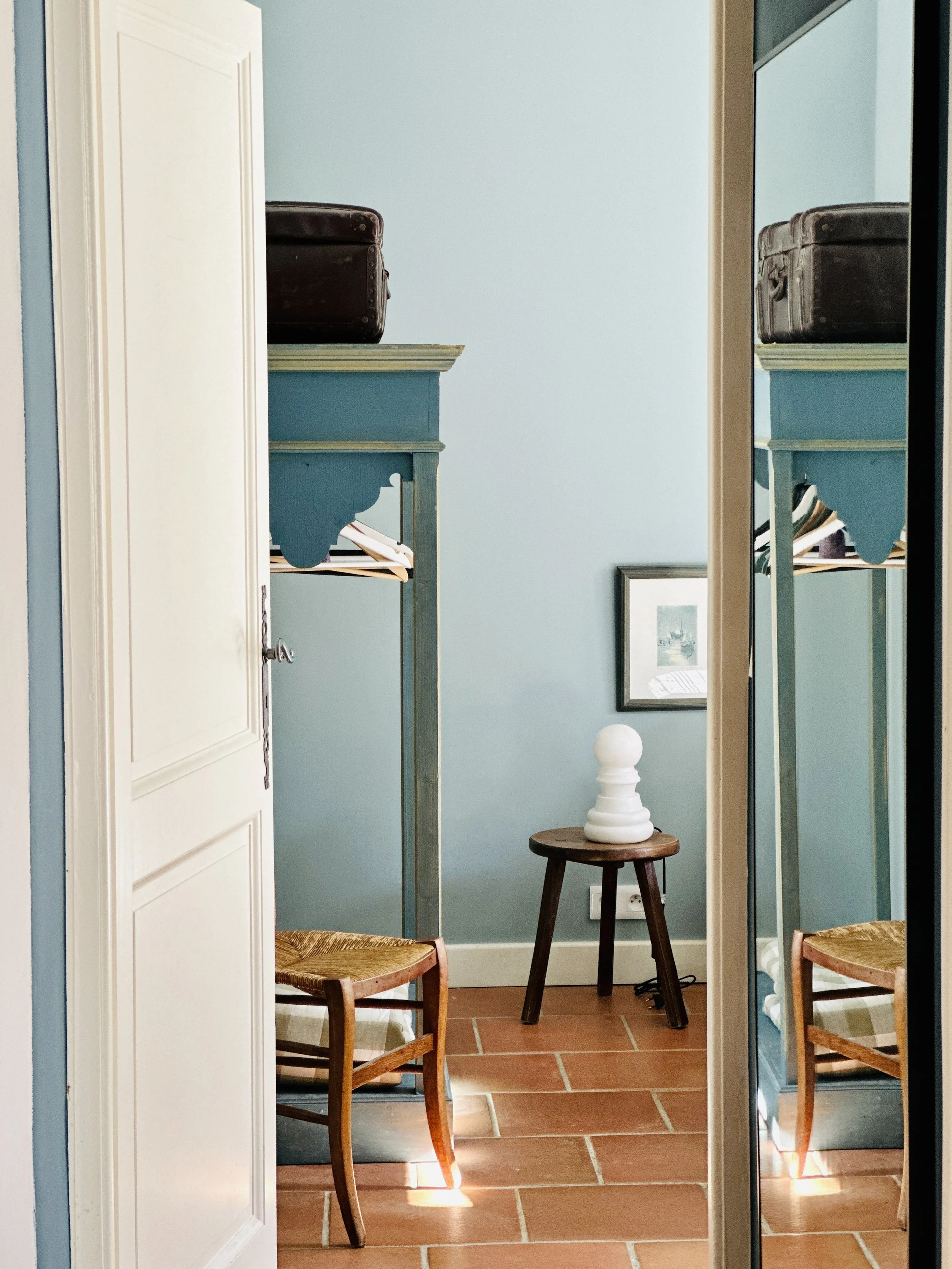 Vue à travers une porte entrouverte sur chambre bleu-gris avec armoire ancienne, tabouret en bois avec lampe sculpturale et miroir mural