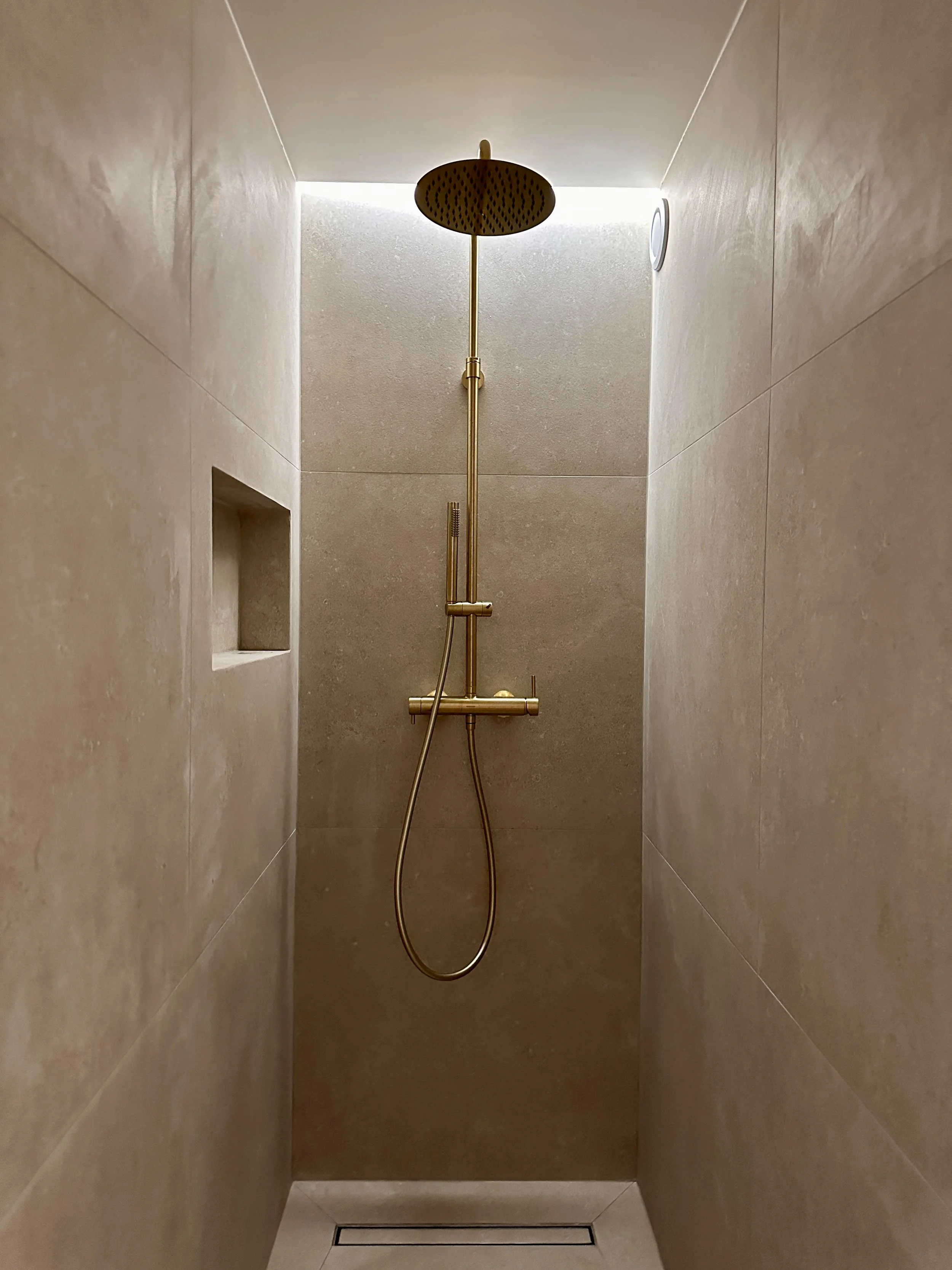Douche à l'italienne en carrelage beige grand format, colonne et pomme de douche en laiton brossé, niche murale et éclairage indirect