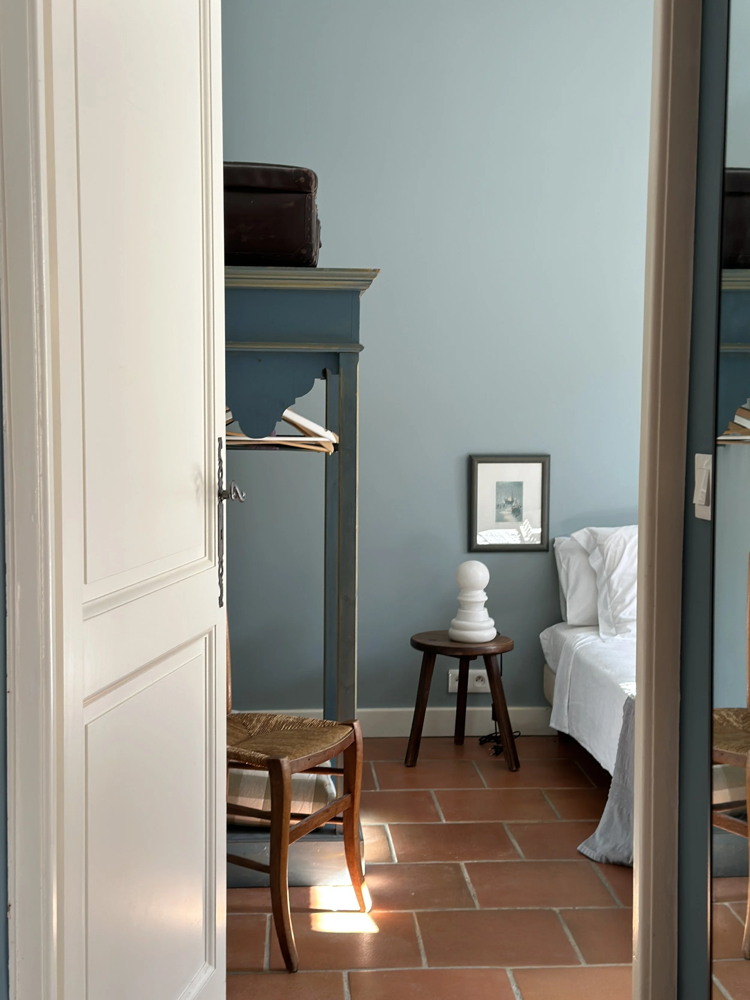 Chambre aux murs bleu-gris avec lit en lin blanc, tabouret en bois avec lampe sculpturale, chaise vintage et sol en tomettes