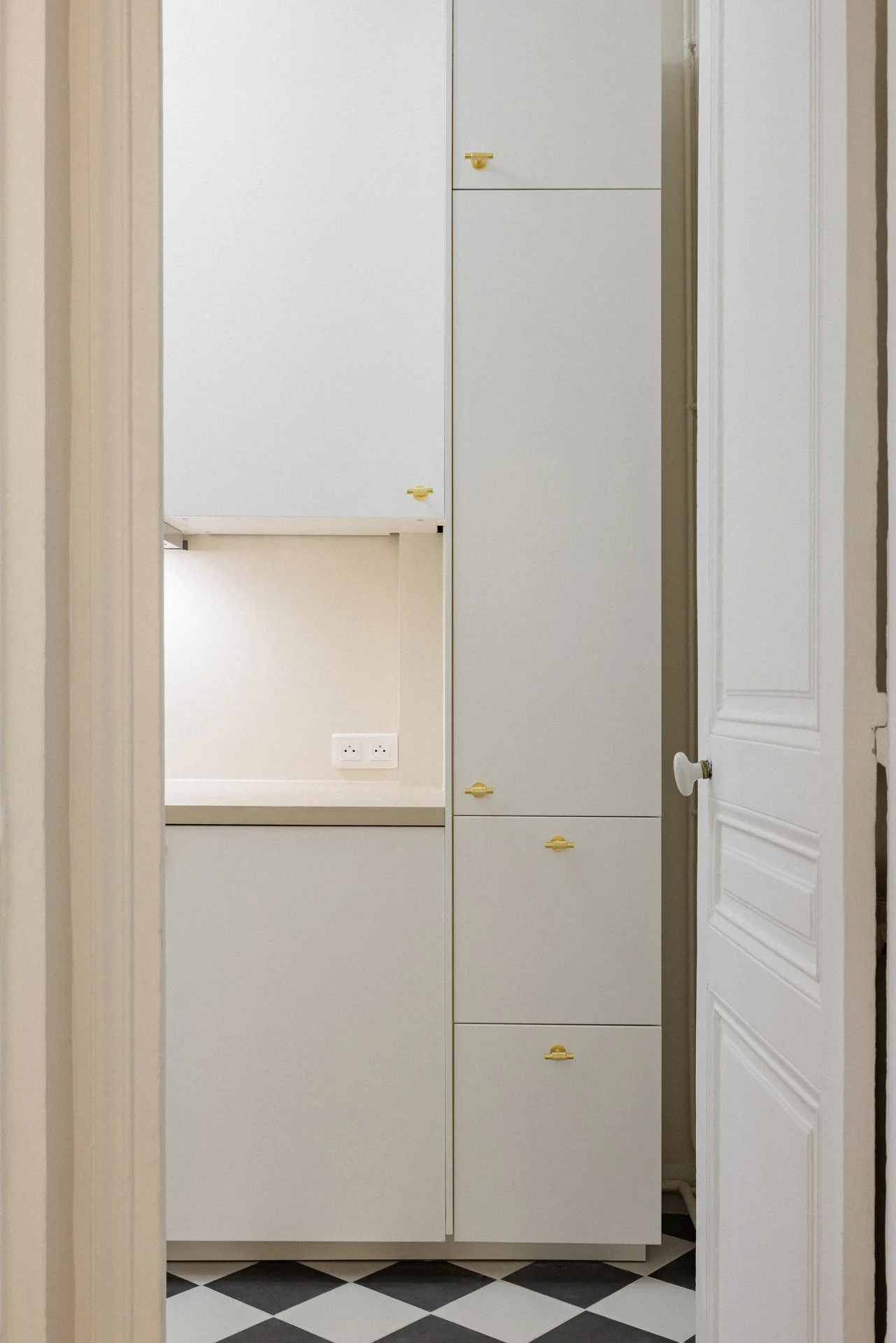 Enfilade de placards sur mesure en encadrement de porte, laiton et sol en damier noir et blanc, avec un renfoncement de rangement dissimulé derrière l'un des vantaux.