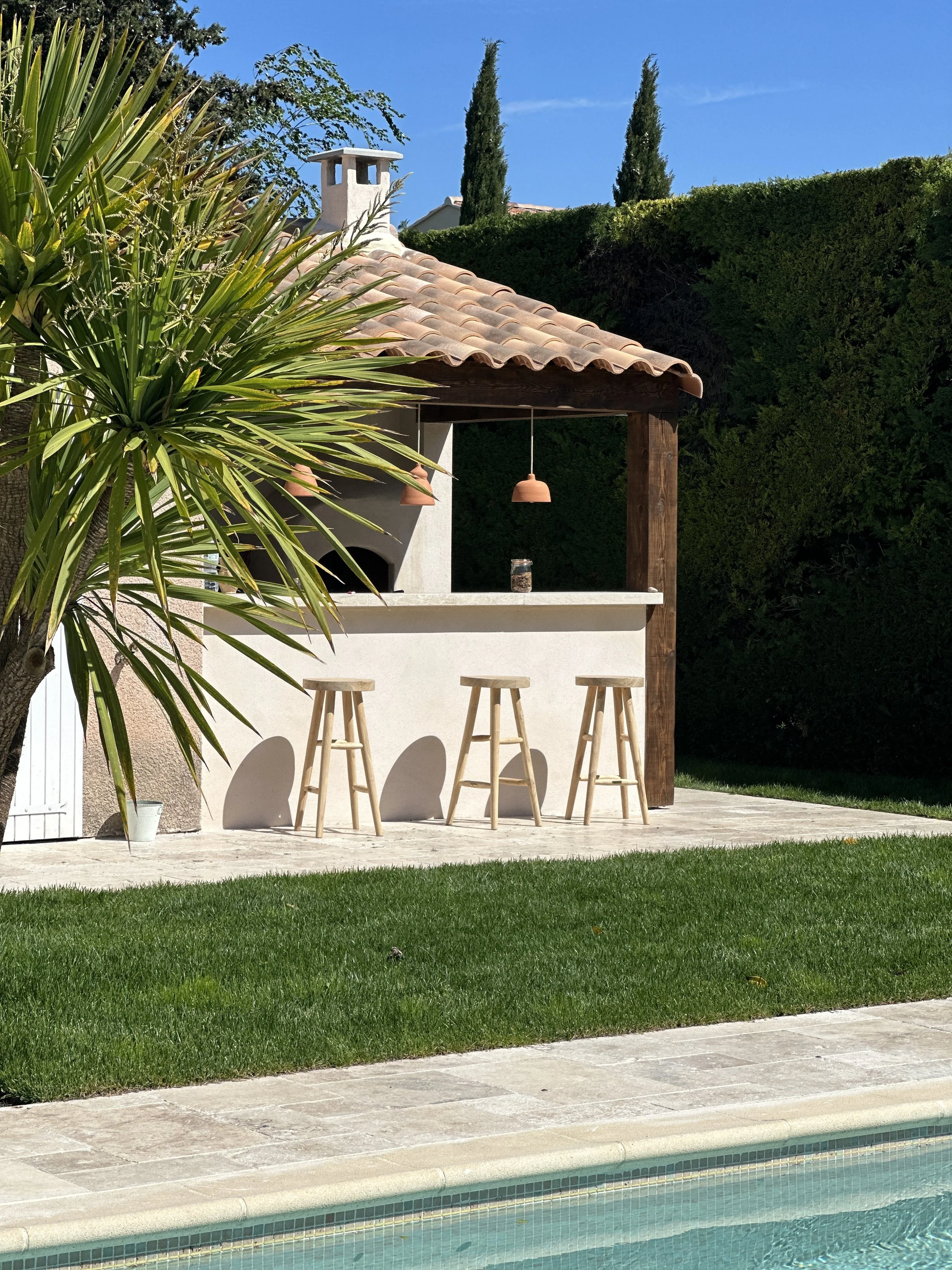 Cuisine d'été en pierre dessinée sur mesure avec son bar et ses tabourets en bois, au bord de la piscine