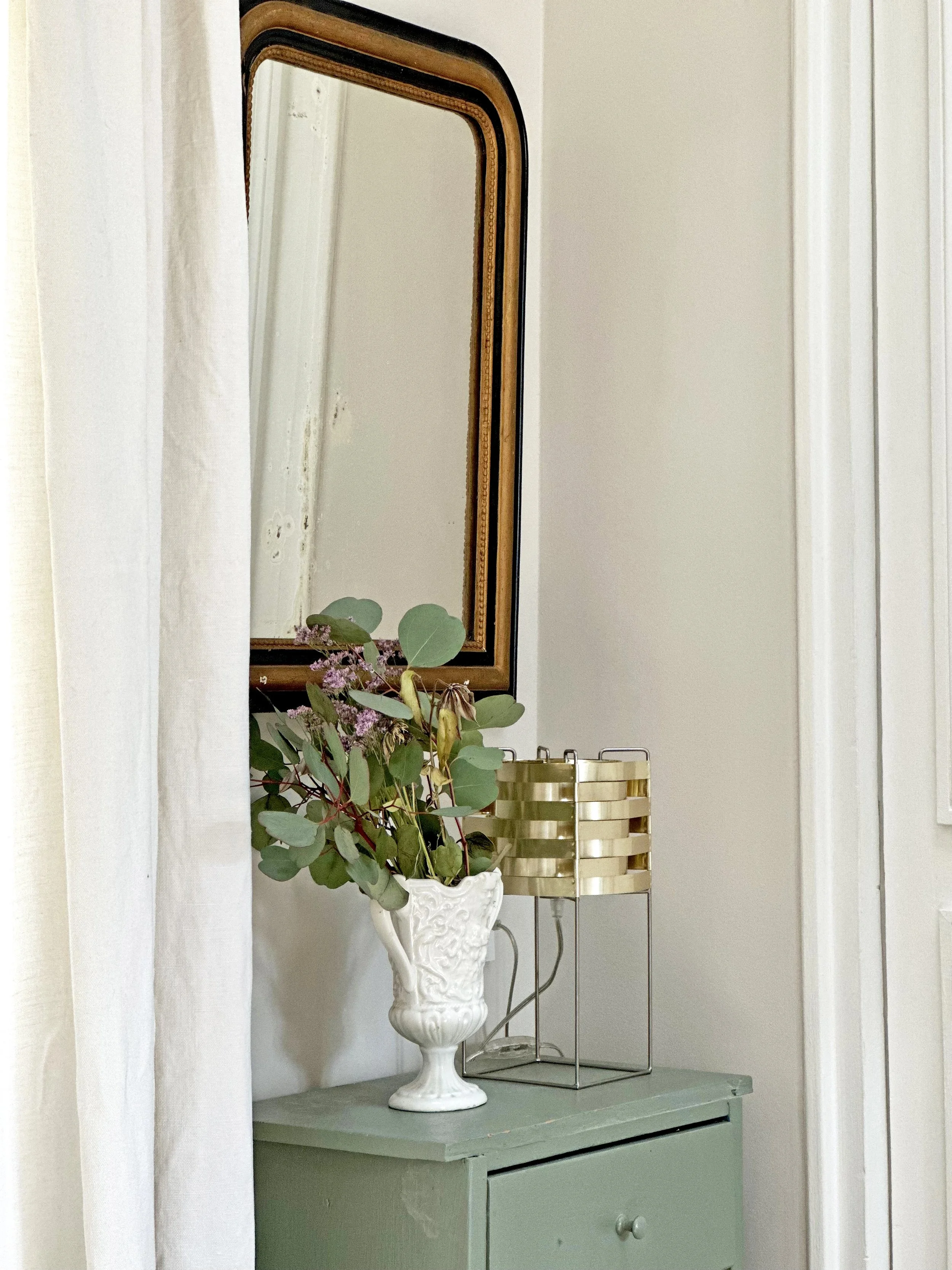 Entrée avec miroir ancien à fronton en bois doré, chevet vert amande, bouquet dans un vase blanc et rideau de lin