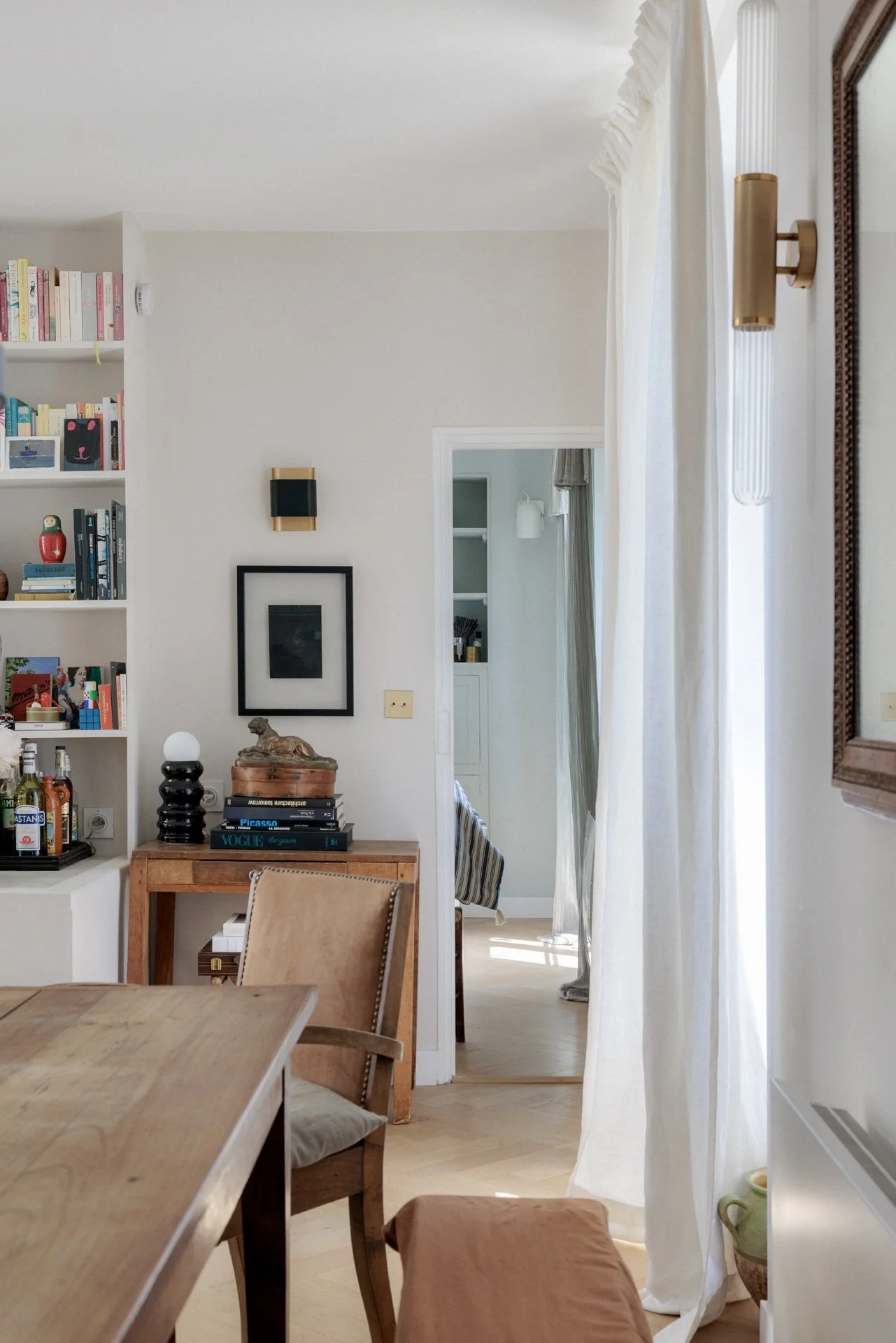 Salle à manger avec bibliothèque, console en bois ancien, livres empilés, chaise en cuir clouté et rideaux blancs devant fenêtre lumineuse