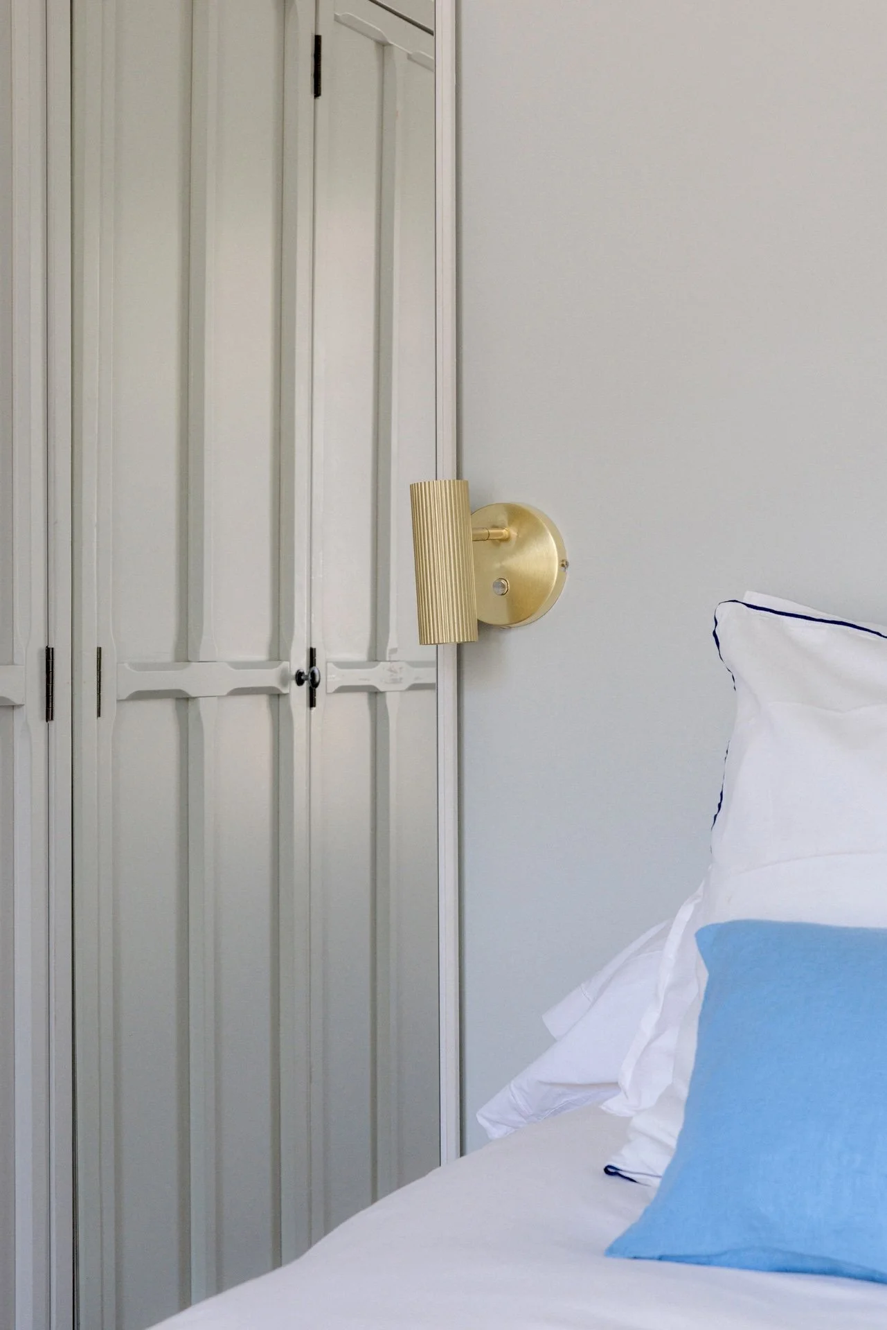 Chambre à coucher avec placard à portes battantes bleu gris, applique murale dorée et lit avec linge blanc à liseré marine et coussin bleu