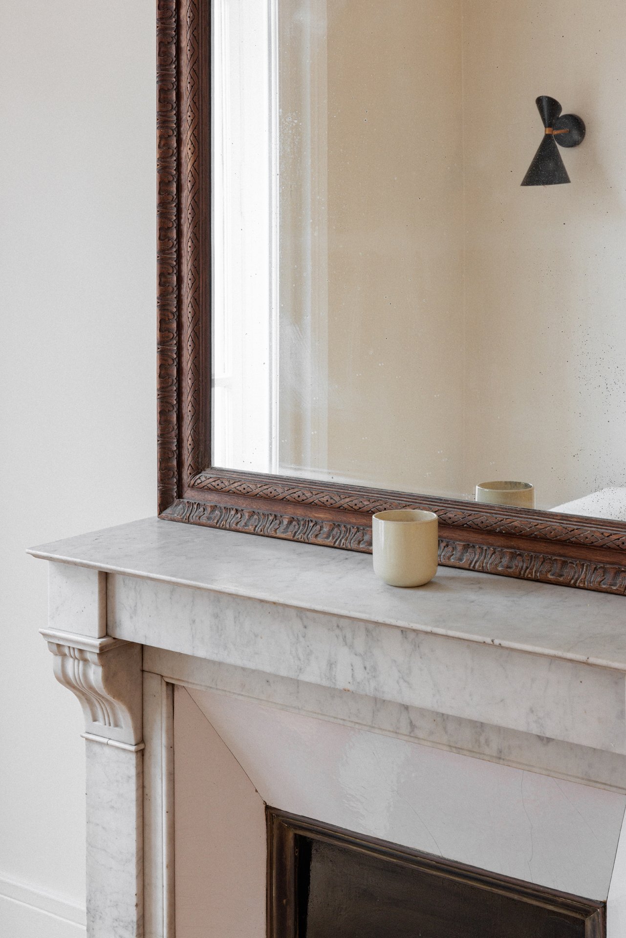 Détail de cheminée en marbre de Carrare avec miroir ancien à cadre en bois sculpté et tasse en grès émaillé, applique noire à double cône visible dans le reflet.