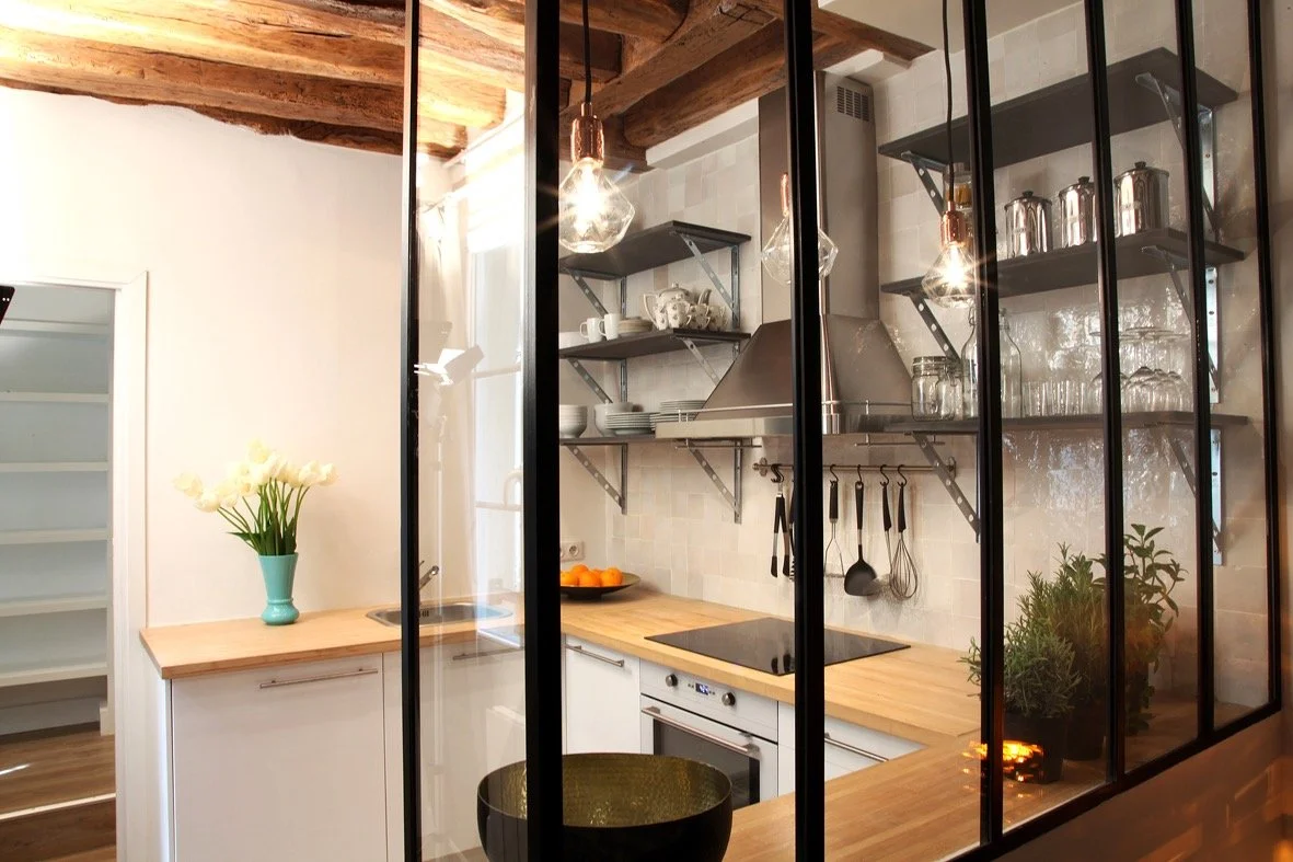 Cuisine semi-ouverte derrière une verrière métallique sur mesure, plan de travail en bois, crédence en zellige et poutres apparentes