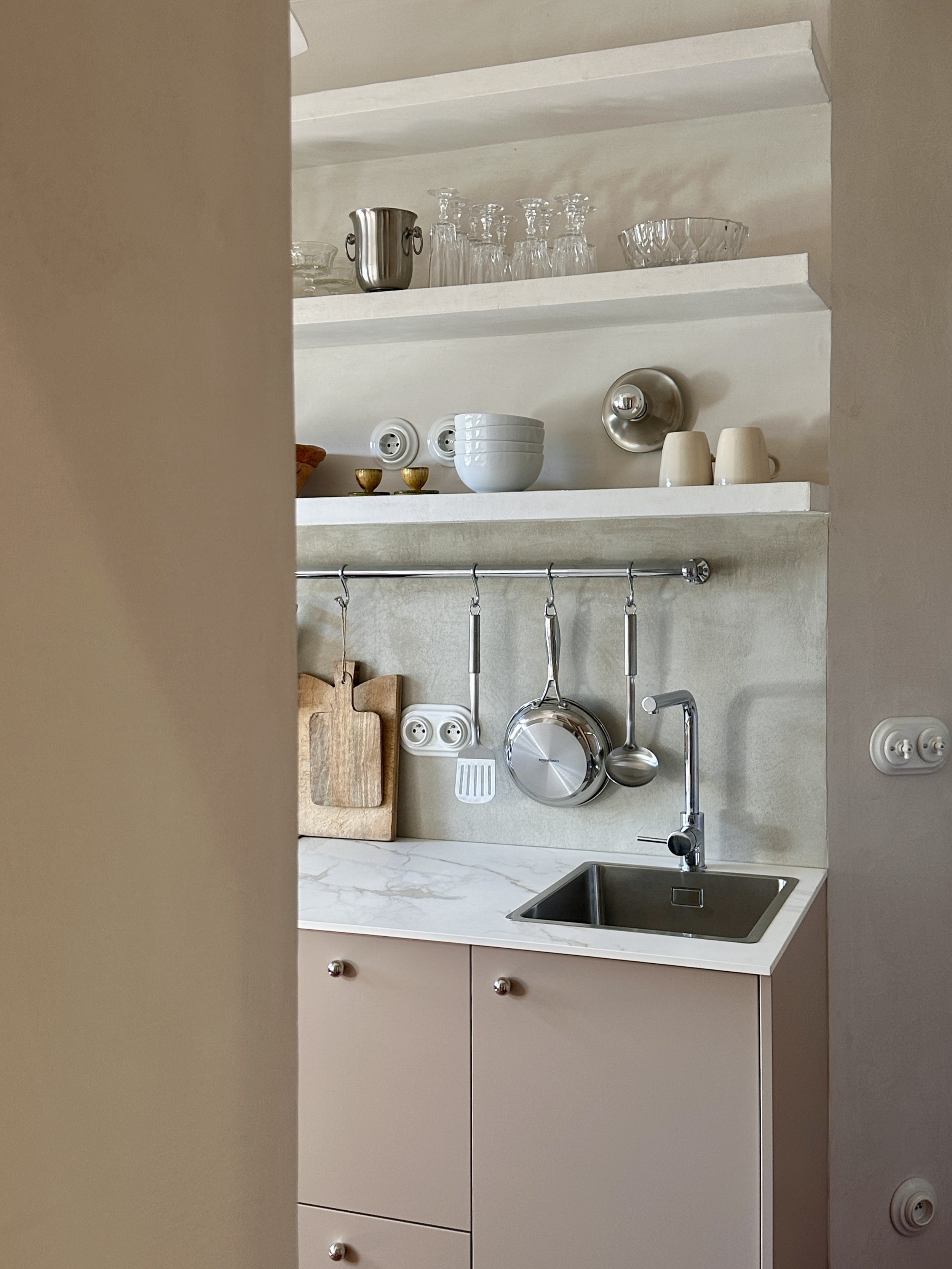 Vue à travers une porte sur cuisine rose poudré avec étagères ouvertes, ustensiles suspendus et évier sous plan de travail en marbre